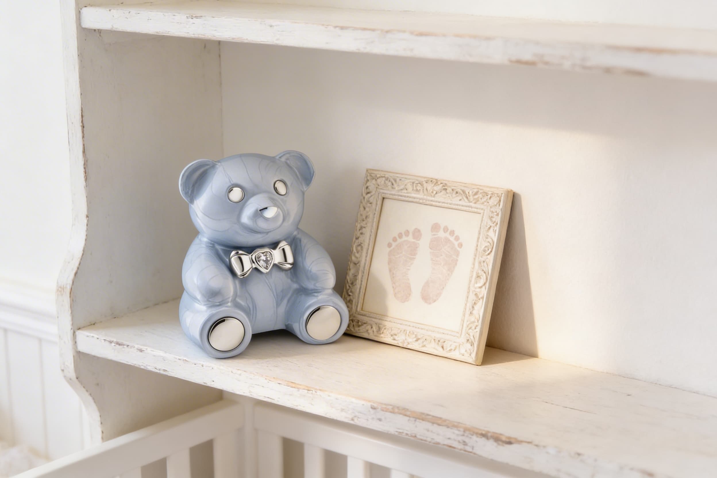 A soft blue ceramic teddy bear cremation urn with a silver bow tie displayed on a white nursery shelf beside a framed set of baby footprints