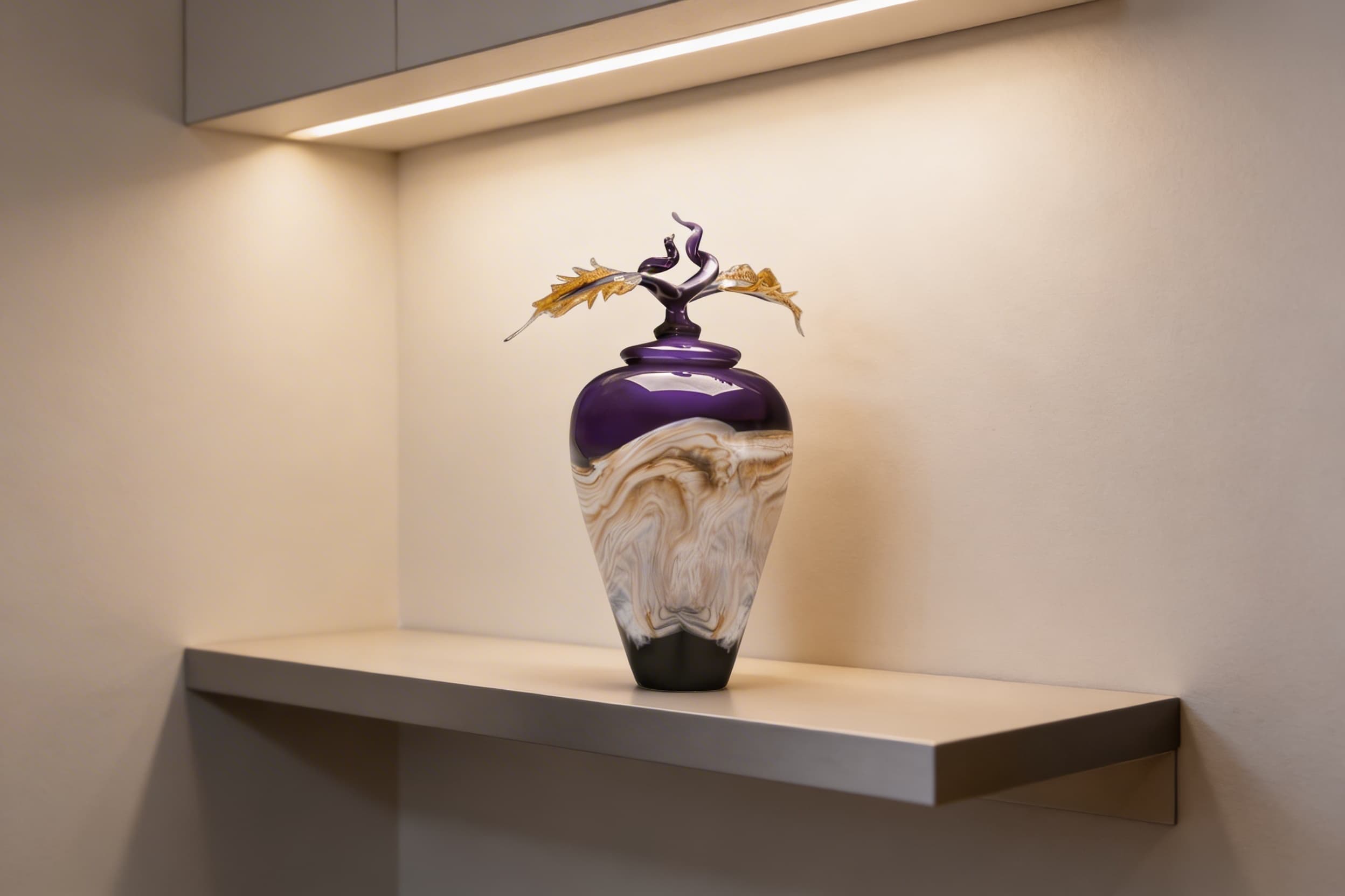 A handblown art glass cremation urn in swirled purple, gold, and white with a sculptural glass leaf topper displayed on a modern floating shelf under accent lighting