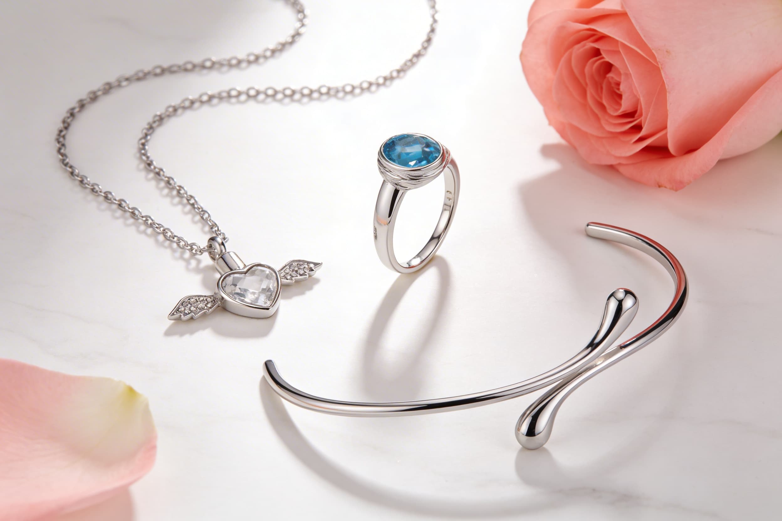 Three pieces of sterling silver cremation jewelry displayed on a marble surface beside a pink rose, including a winged heart pendant necklace, a blue topaz memorial ring, and an open cuff bracelet