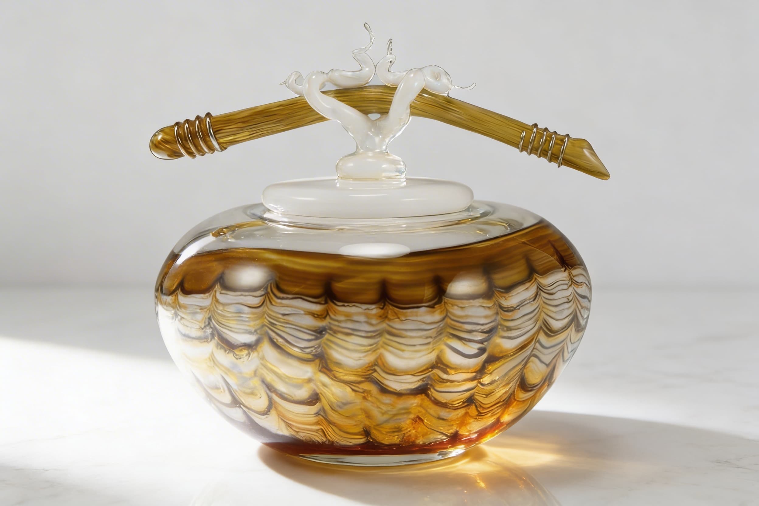 A handblown glass art cremation keepsake with swirled amber and white patterns and a sculptural glass topper, displayed on a marble surface in natural light