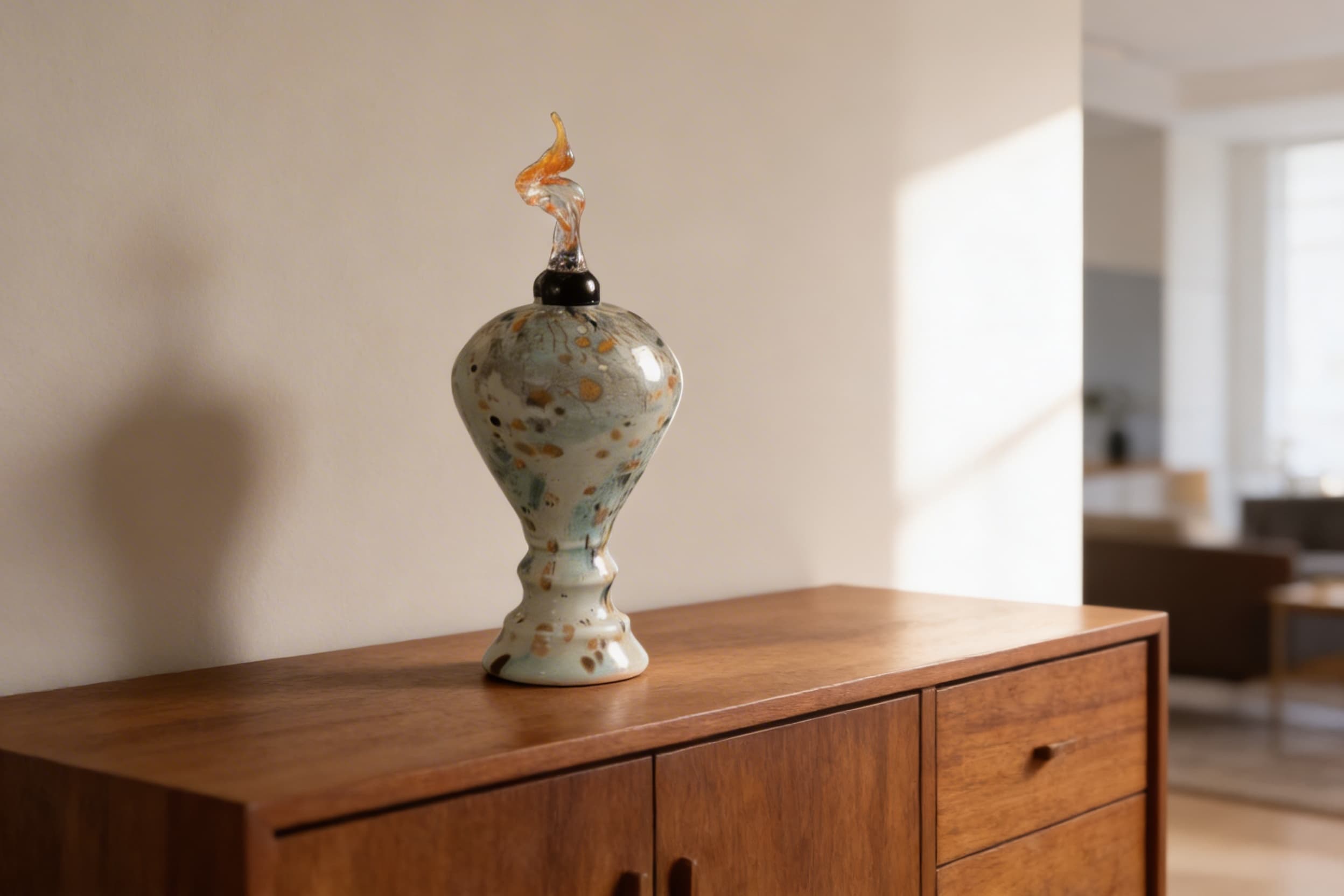A handblown art glass cremation urn with speckled sage green and amber tones and a sculptural flame-shaped glass topper displayed on a mid-century walnut credenza