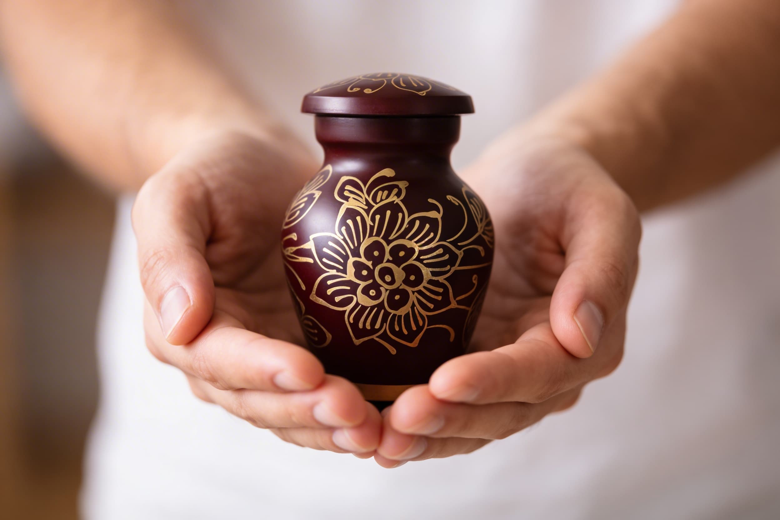 A person holding a small burgundy keepsake cremation urn with a hand-painted gold floral mandala design cupped gently in both hands