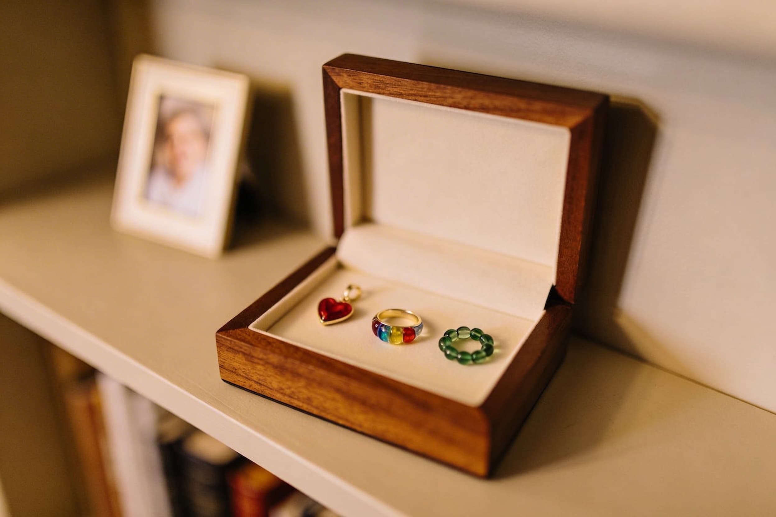 A warmly lit wooden display shelf with several cremation jewelry pieces arranged in an open shadow box -- pendant, ring, and bracelet bead -- with a small framed photo nearby