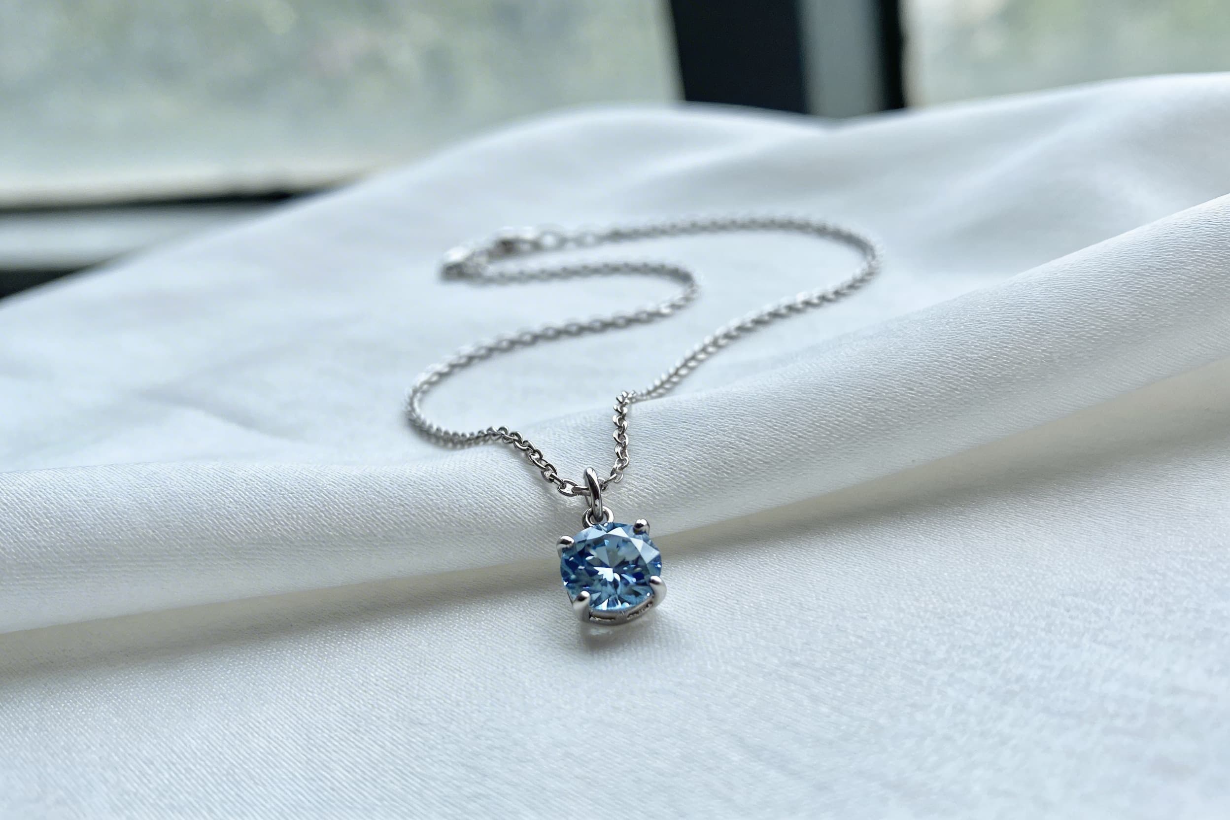 A close-up of a small blue cremation diamond pendant resting on a piece of white silk fabric in natural window light