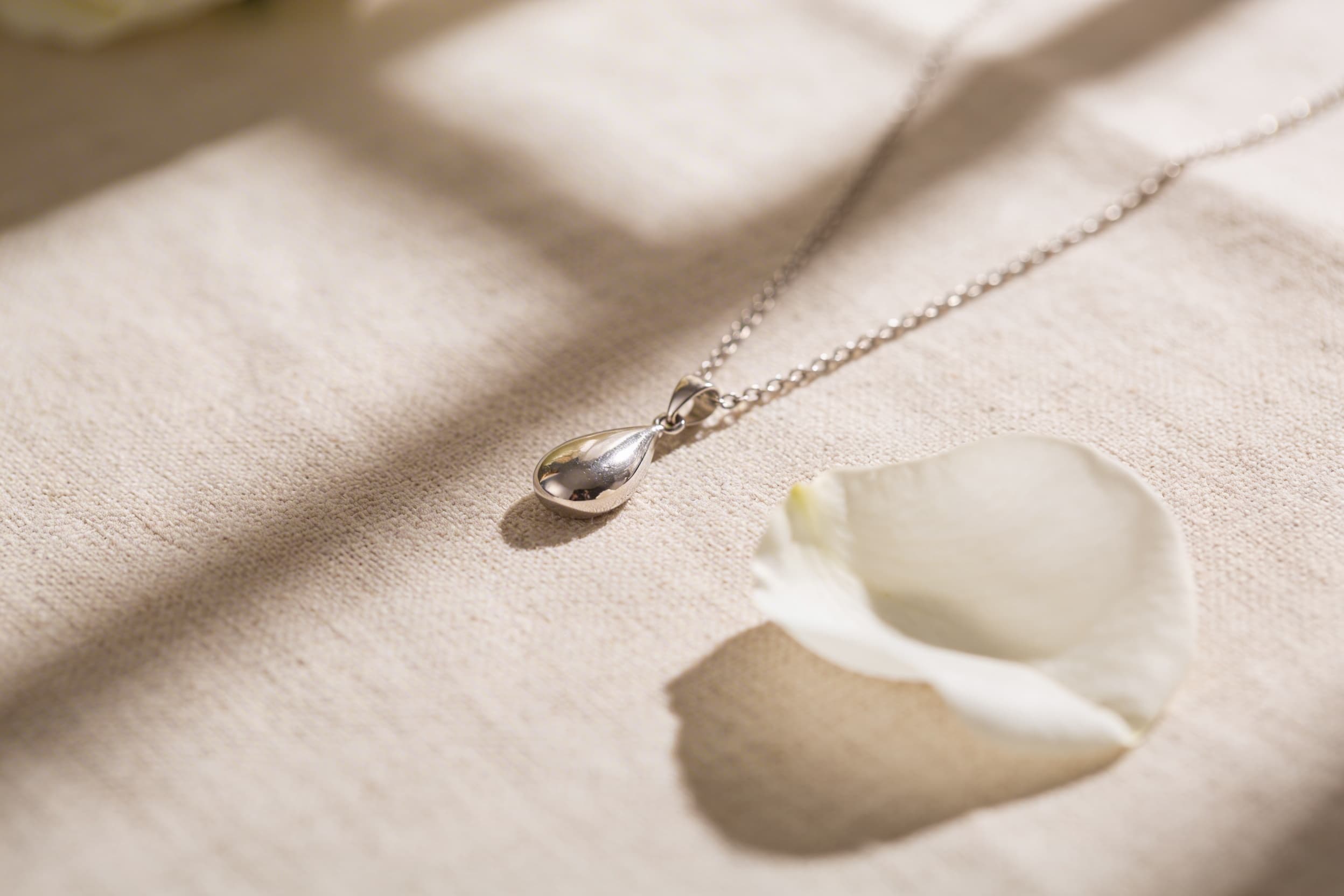 A delicate silver cremation jewelry pendant on a linen surface with soft natural light and a single white flower beside it
