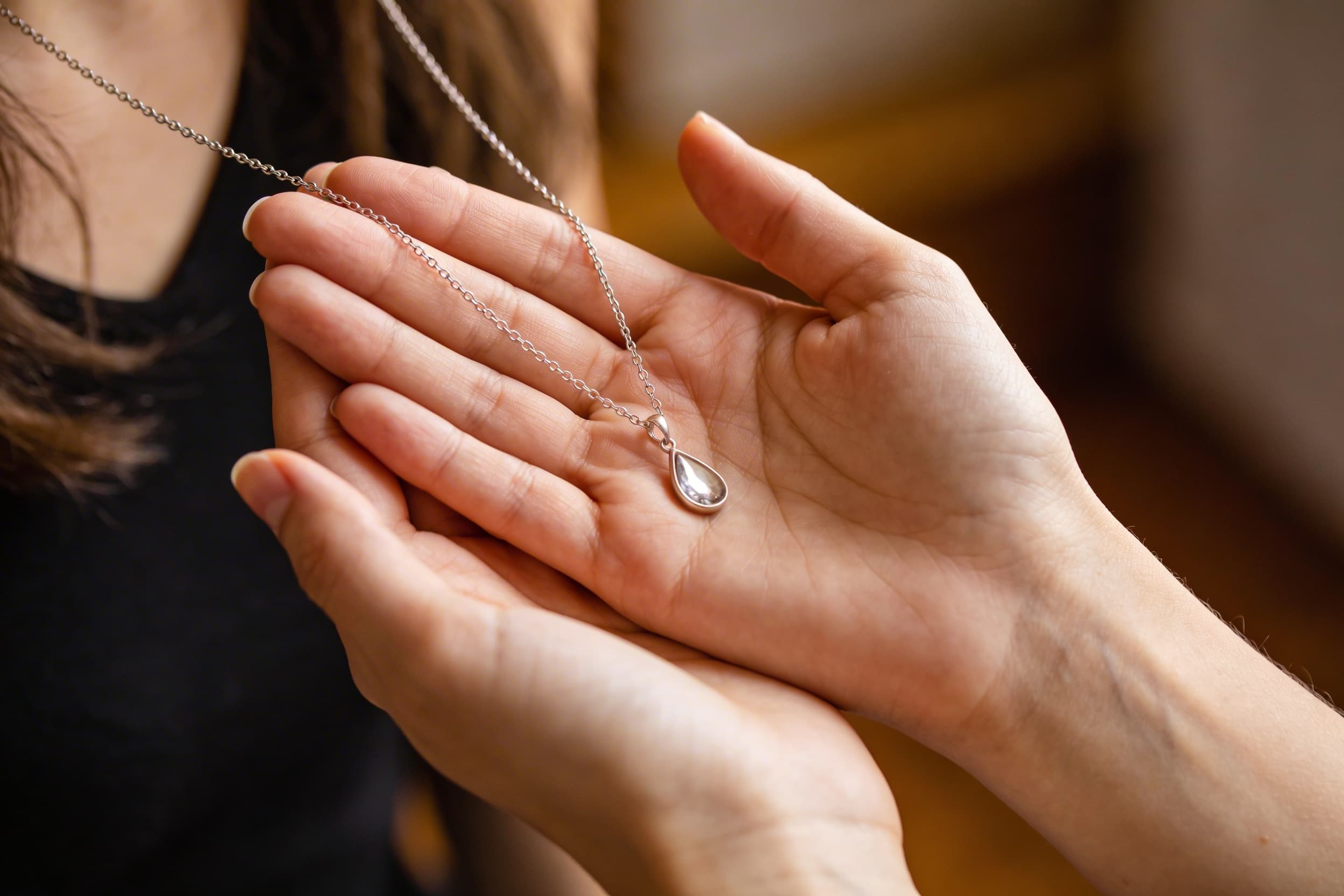 A woman's hand gently holding a small teardrop silver cremation pendant, soft natural light and blurred background