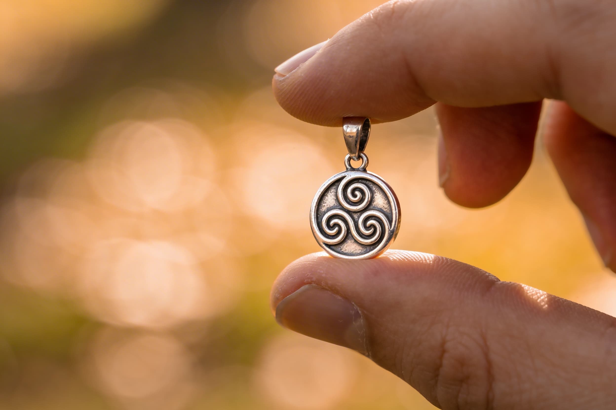 A small triskelion triple spiral cremation jewelry pendant held between a person's fingers, sterling silver, warm bokeh background, memorial photography