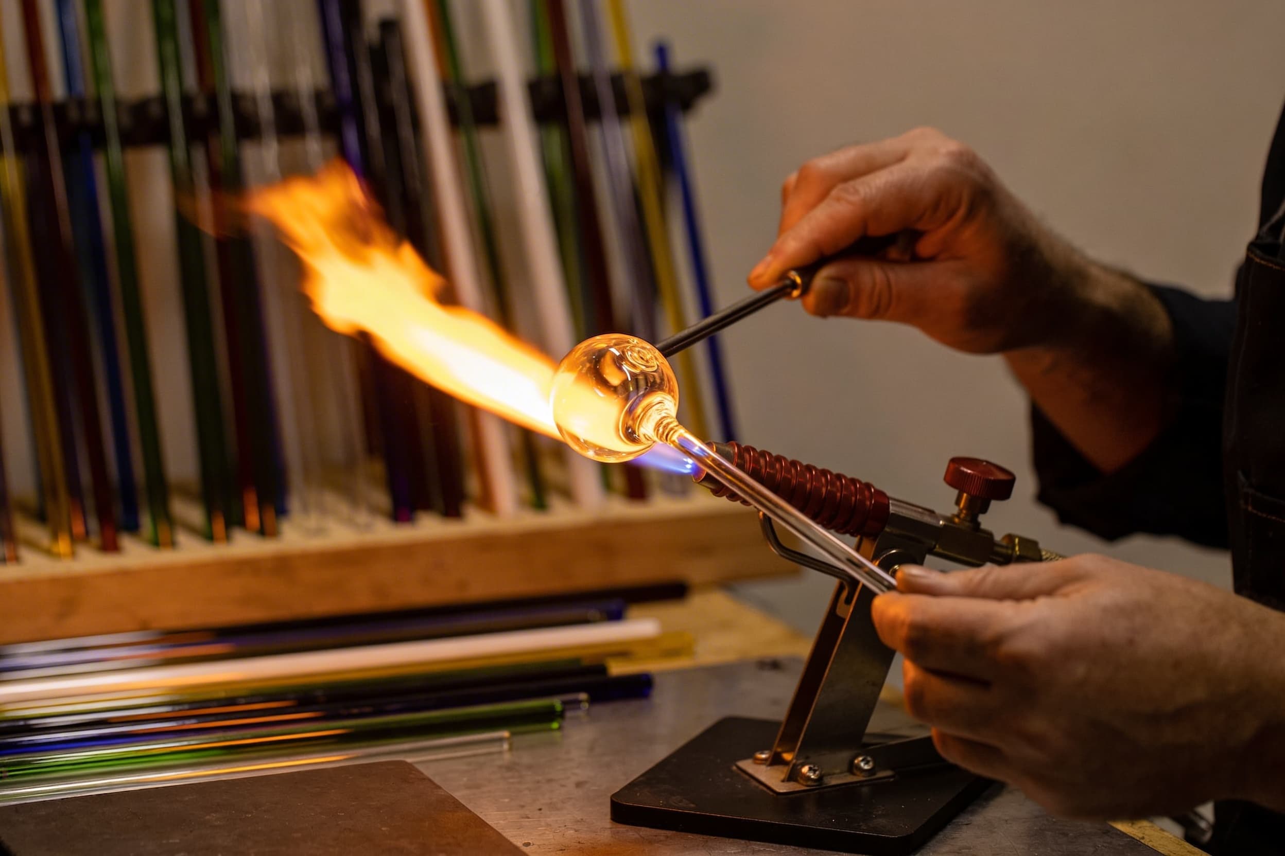 A glass artisan using a handheld torch to shape molten borosilicate glass in a warm studio workshop, surrounded by colored glass rods