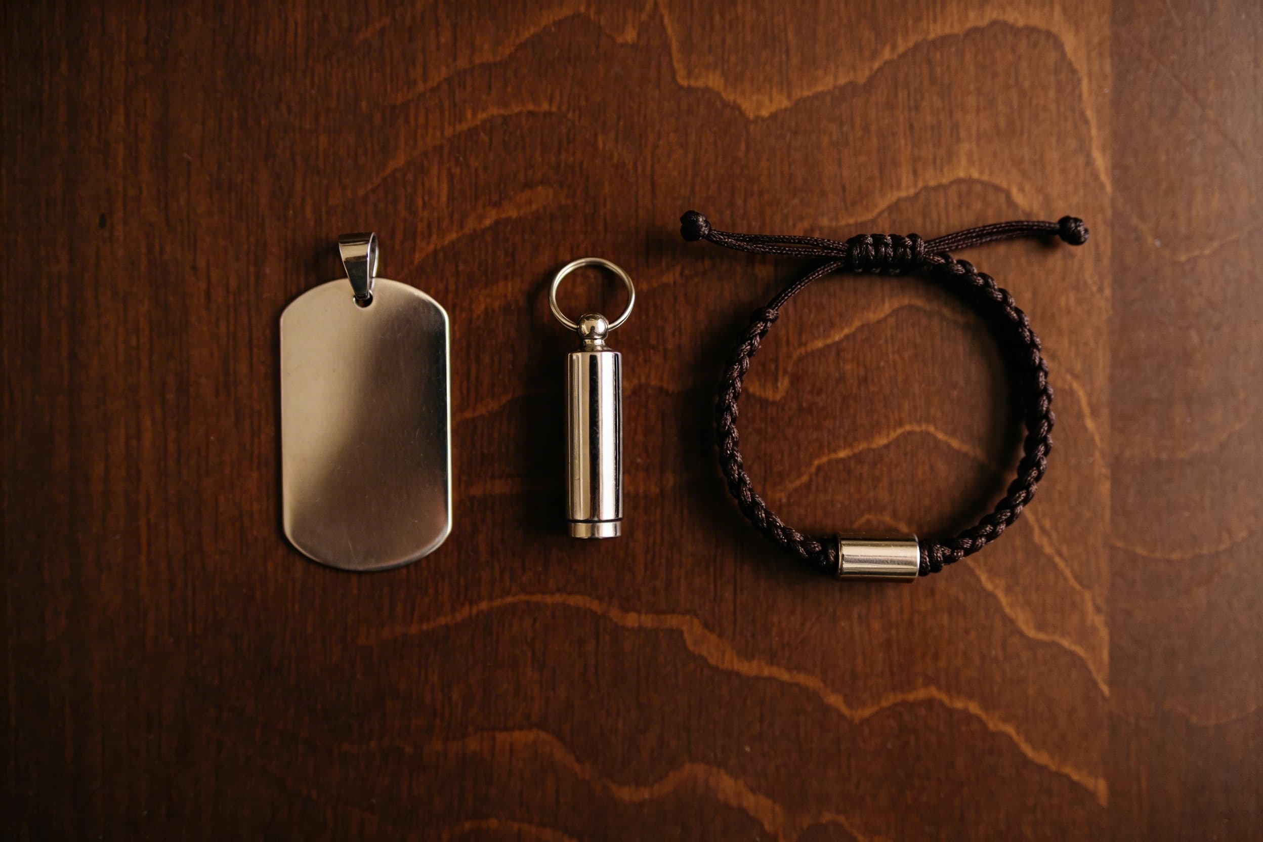A dark wood surface with a stainless steel dog tag cremation pendant, a simple keychain urn, and a cord bracelet with a metal bead arranged neatly together, soft overhead light
