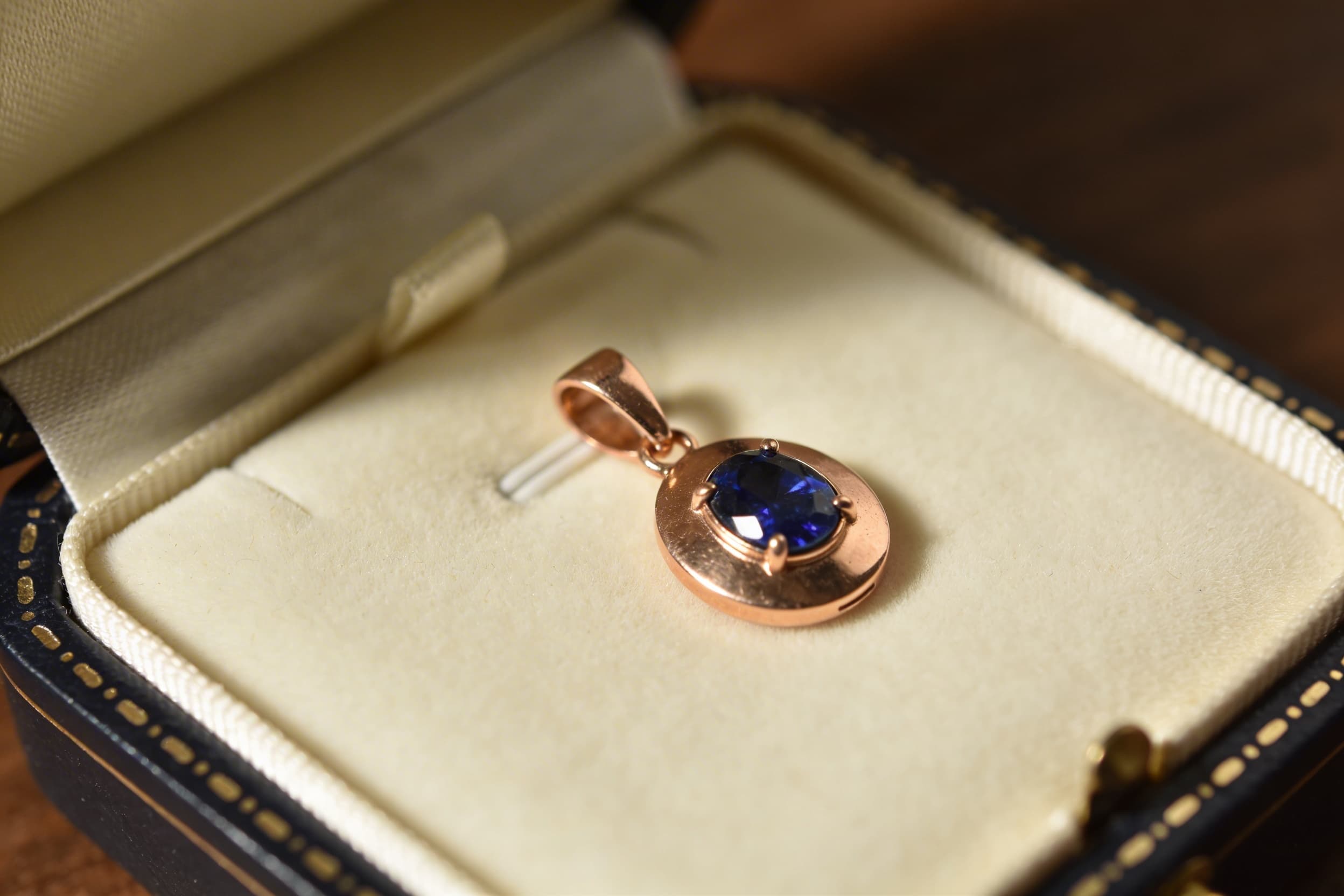 A close-up of a birthstone cremation pendant in rose gold showing a deep blue sapphire accent stone, displayed in an open jewelry box with cream interior