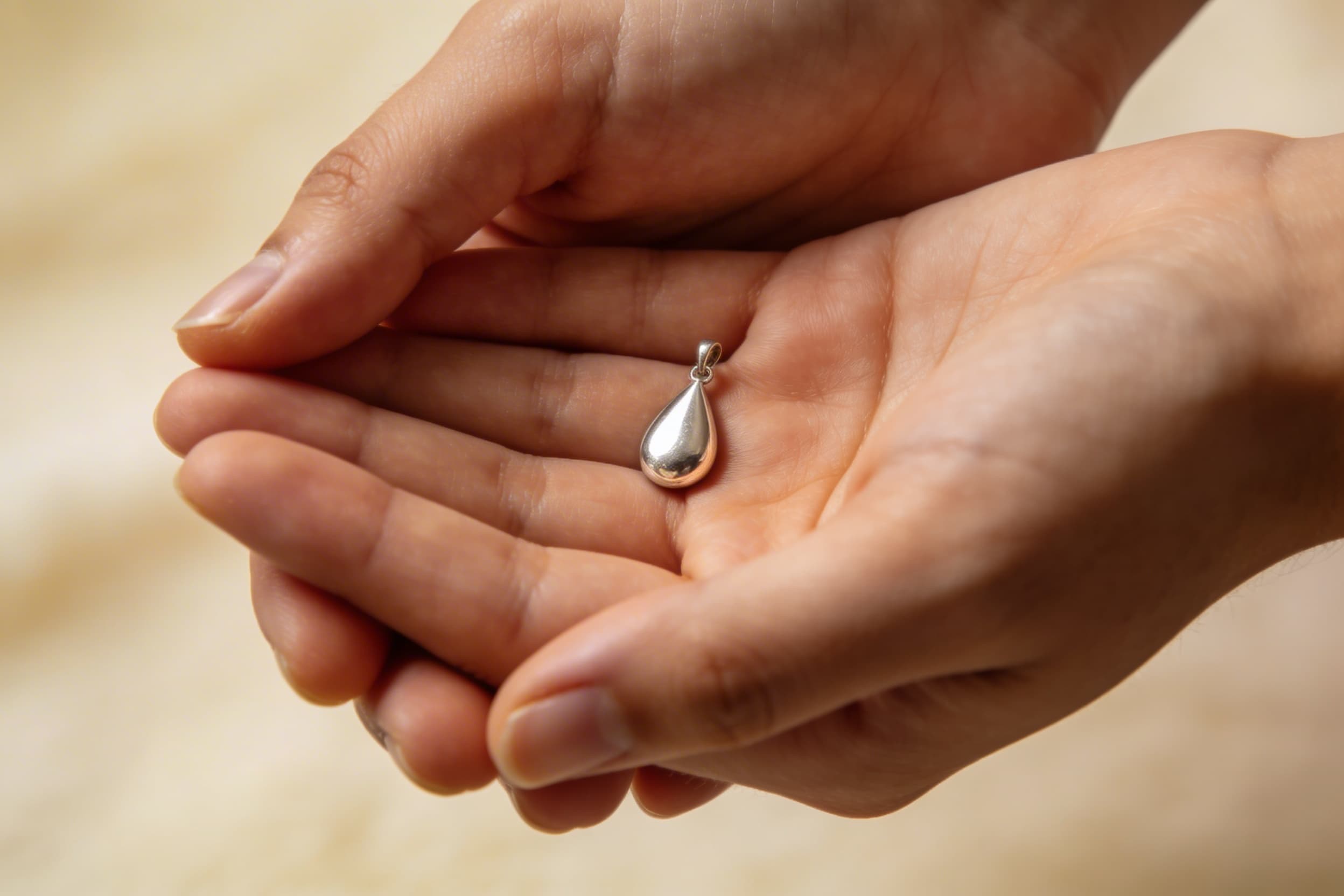 Close-up of a person's hand holding a small silver teardrop cremation pendant, soft bokeh background, warm indoor light