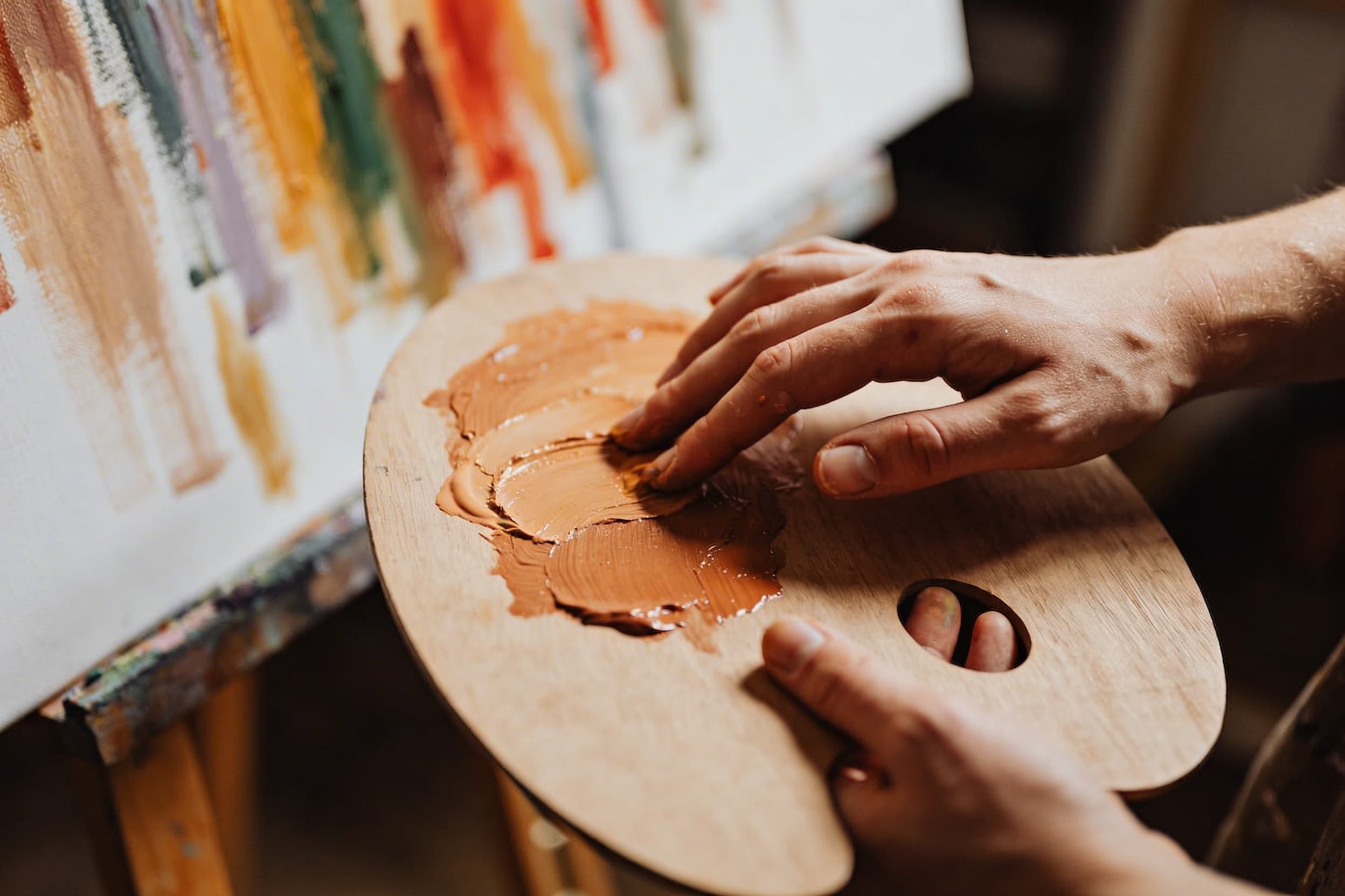 An artist's hands carefully blending paint medium on a wooden palette, with a memorial canvas in soft focus behind, warm studio lighting