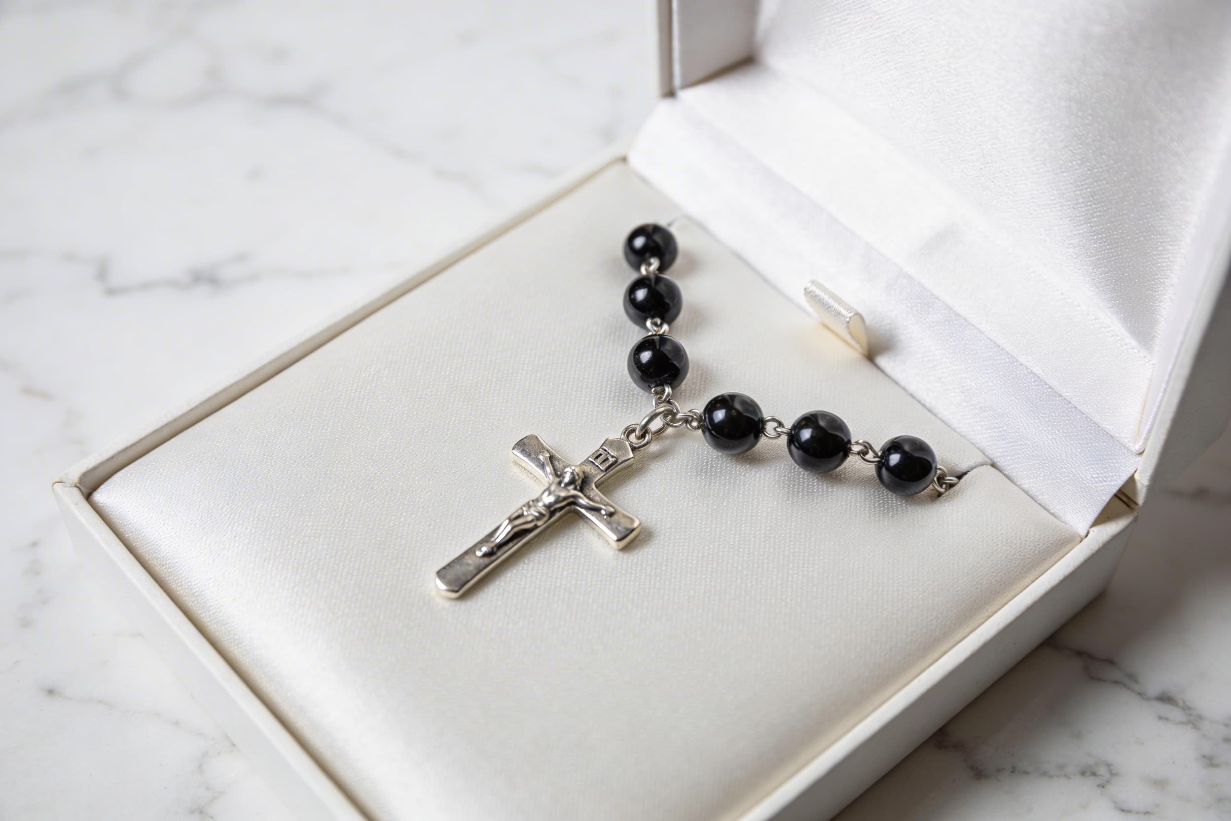 A cremation rosary with dark natural stone beads and a silver crucifix displayed in an open presentation box on white marble