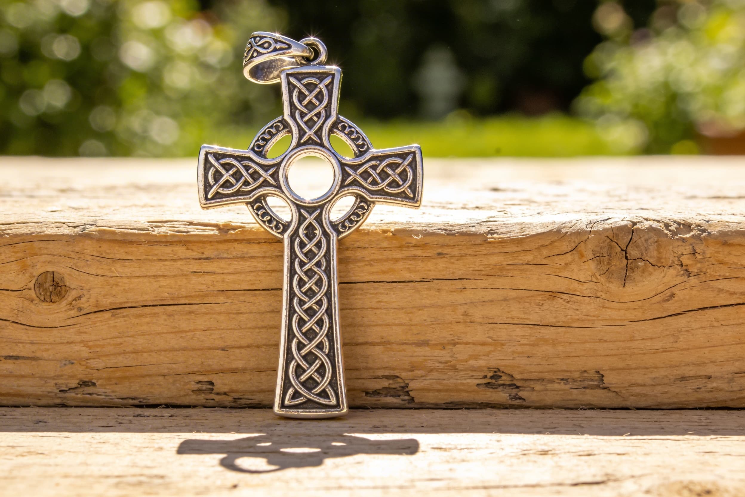 An ornate Celtic cross cremation pendant in sterling silver resting on a weathered wooden surface, soft bokeh green background, warm natural lighting