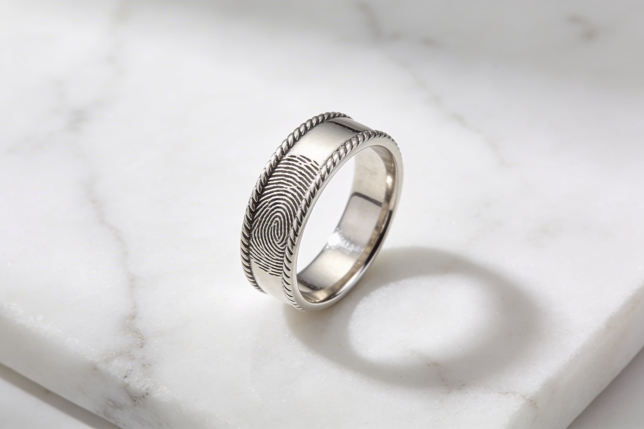 A sterling silver fingerprint memorial ring displayed on a white marble surface, the embossed ridge lines of the print visible along the band