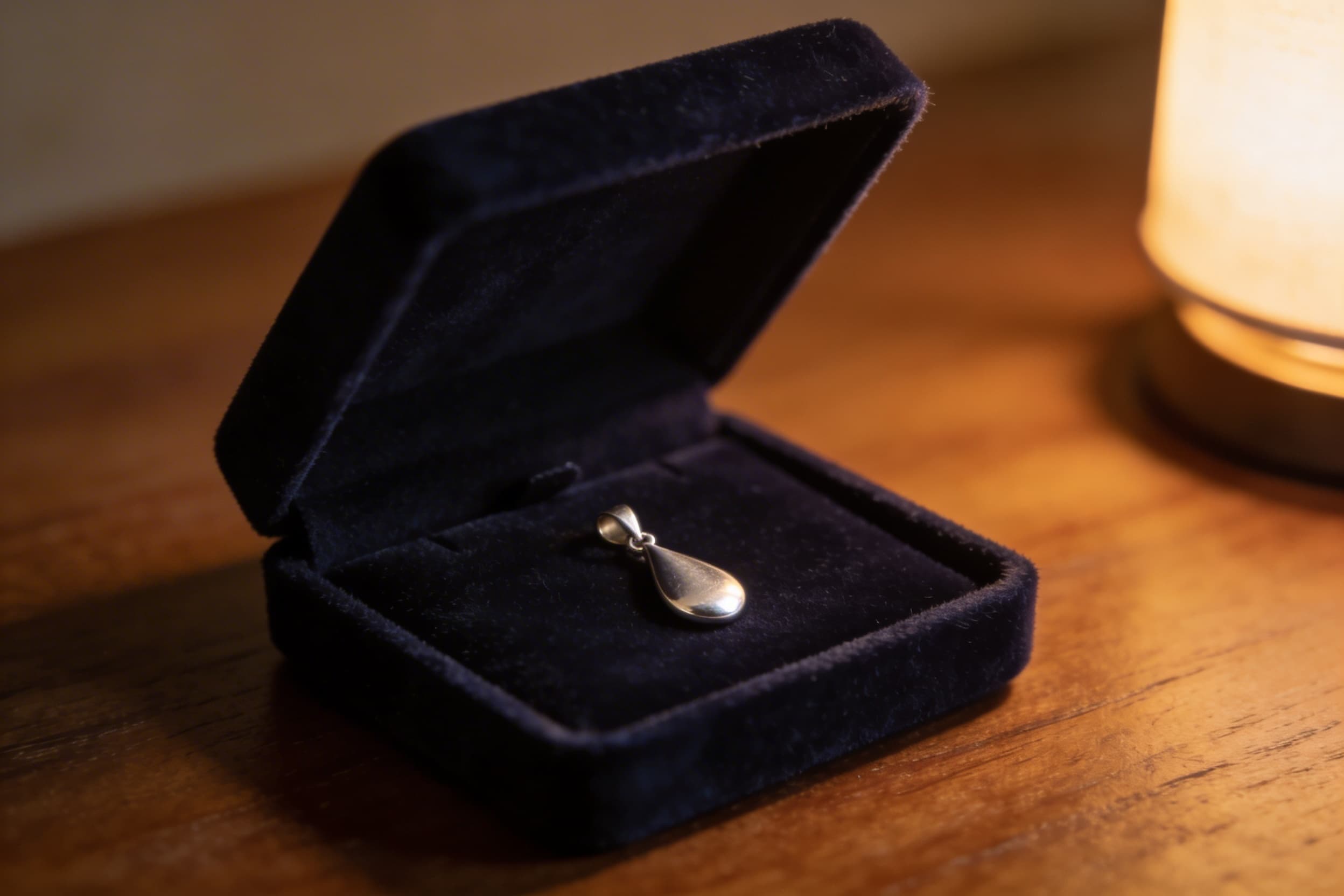A small sterling silver cremation pendant resting in an open velvet jewelry box on a wooden bedside table, soft warm light