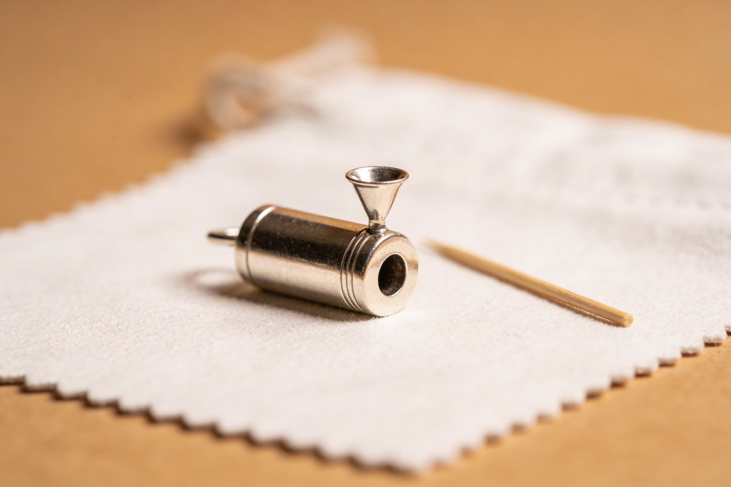Close-up of a small silver cremation pendant with a mini funnel inserted, ashes being carefully guided in with a toothpick