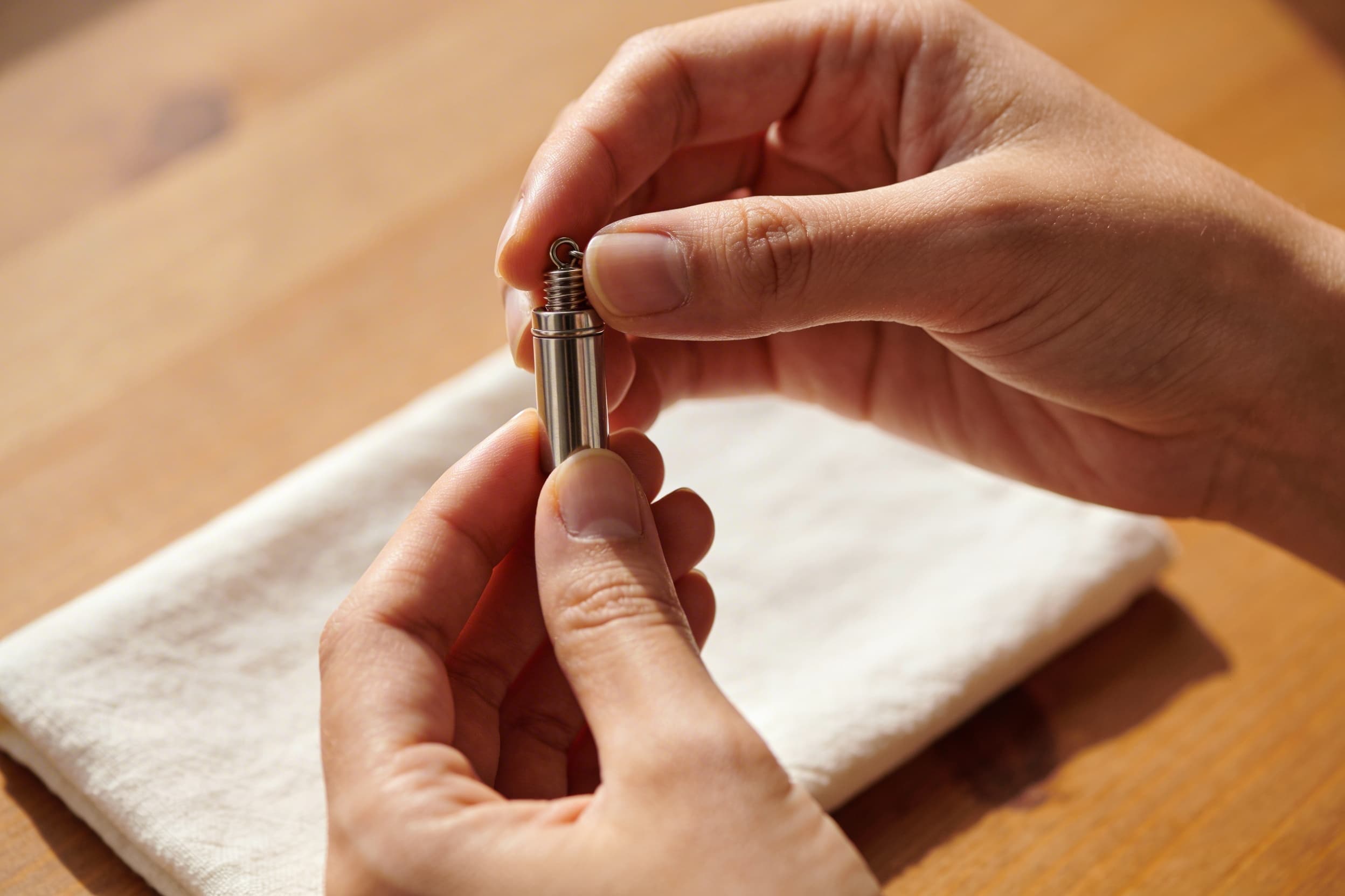 A stainless steel screw-top cremation pendant being gently inspected, with a soft cloth nearby on a wooden table