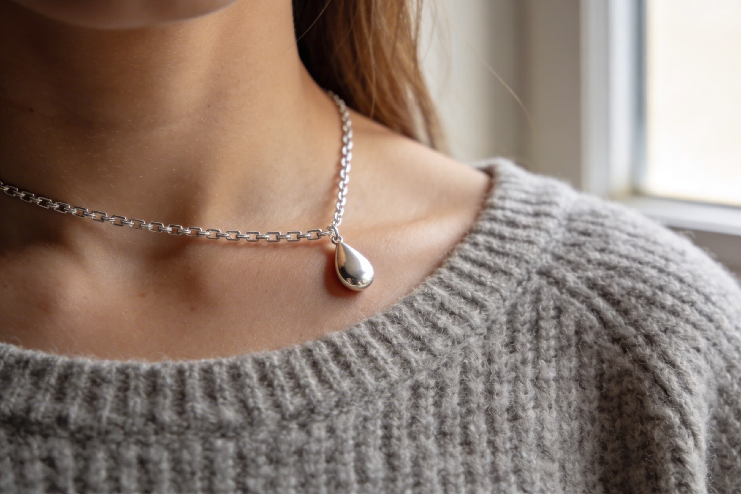 A close-up of a sterling silver cremation pendant necklace resting on a woman's collarbone against a soft gray sweater