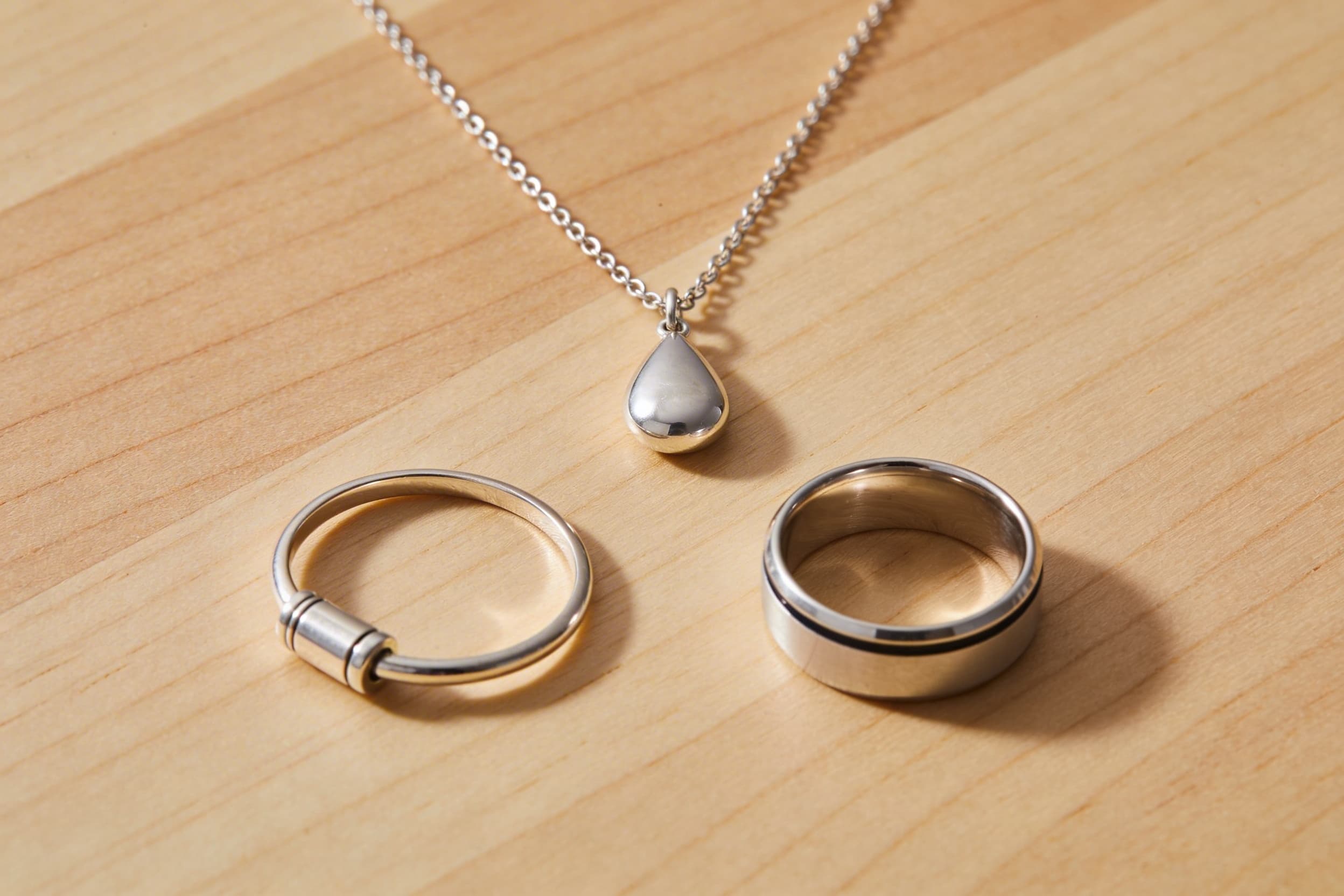 Three pieces of cremation jewelry arranged together โ a pendant necklace, a wristband bracelet, and a band ring โ displayed on a wooden surface