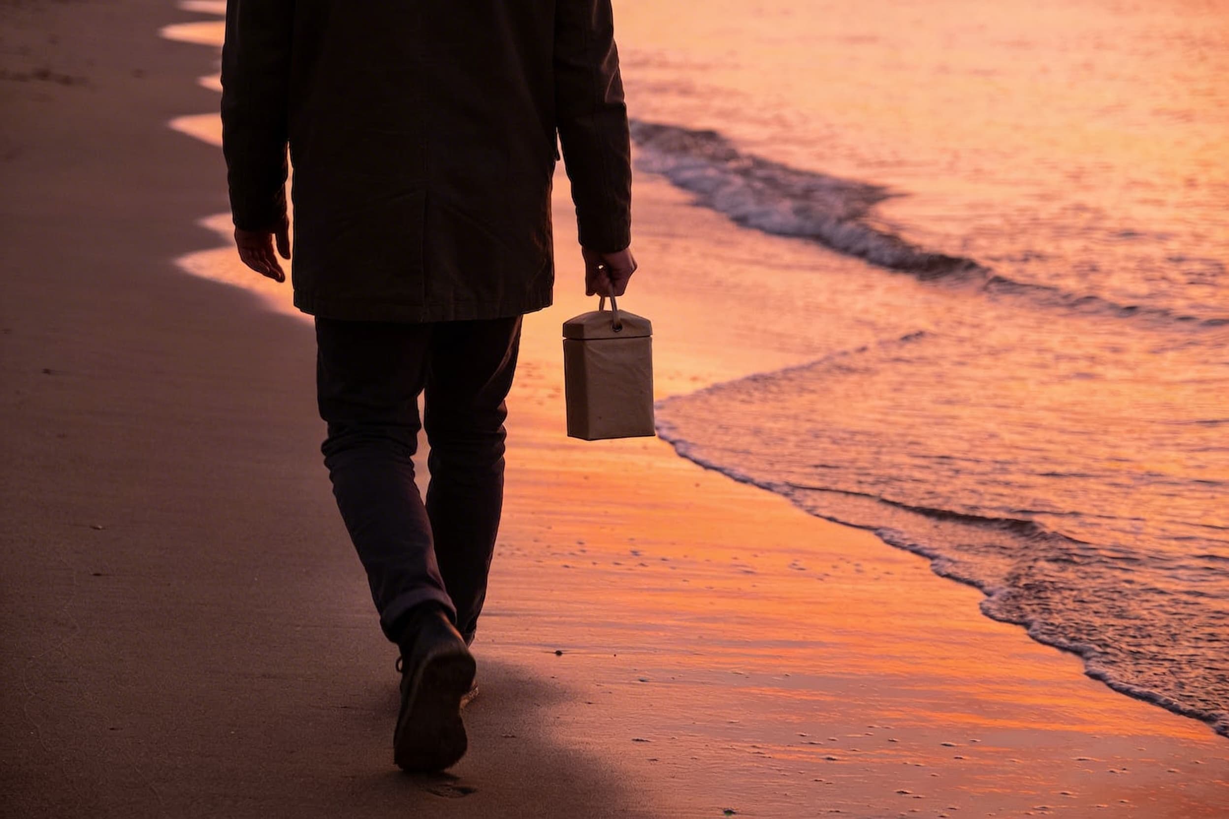 A person walking alone on a peaceful beach at sunset carrying a small biodegradable pet urn