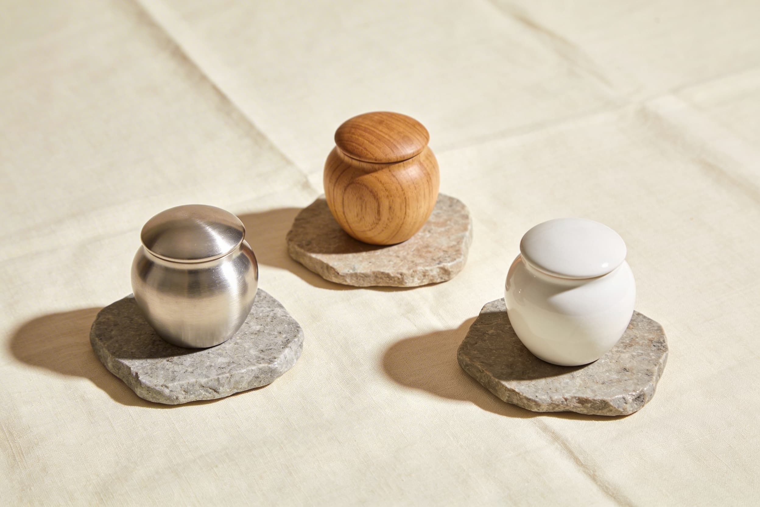 Three small pet keepsake urns in brushed silver, natural wood, and white ceramic on stone coasters