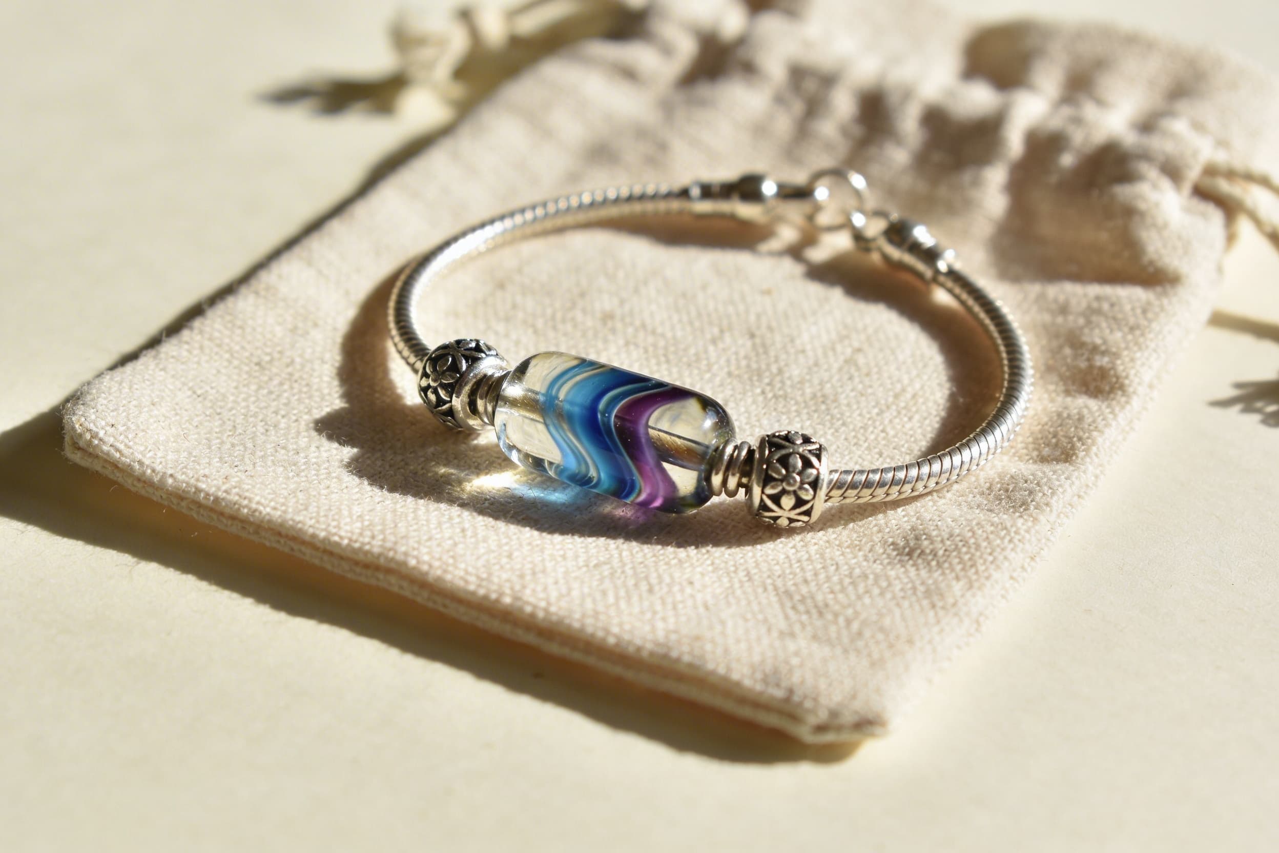 A delicate charm bracelet with a colorful hand-blown glass cremation bead alongside silver charms on natural linen fabric