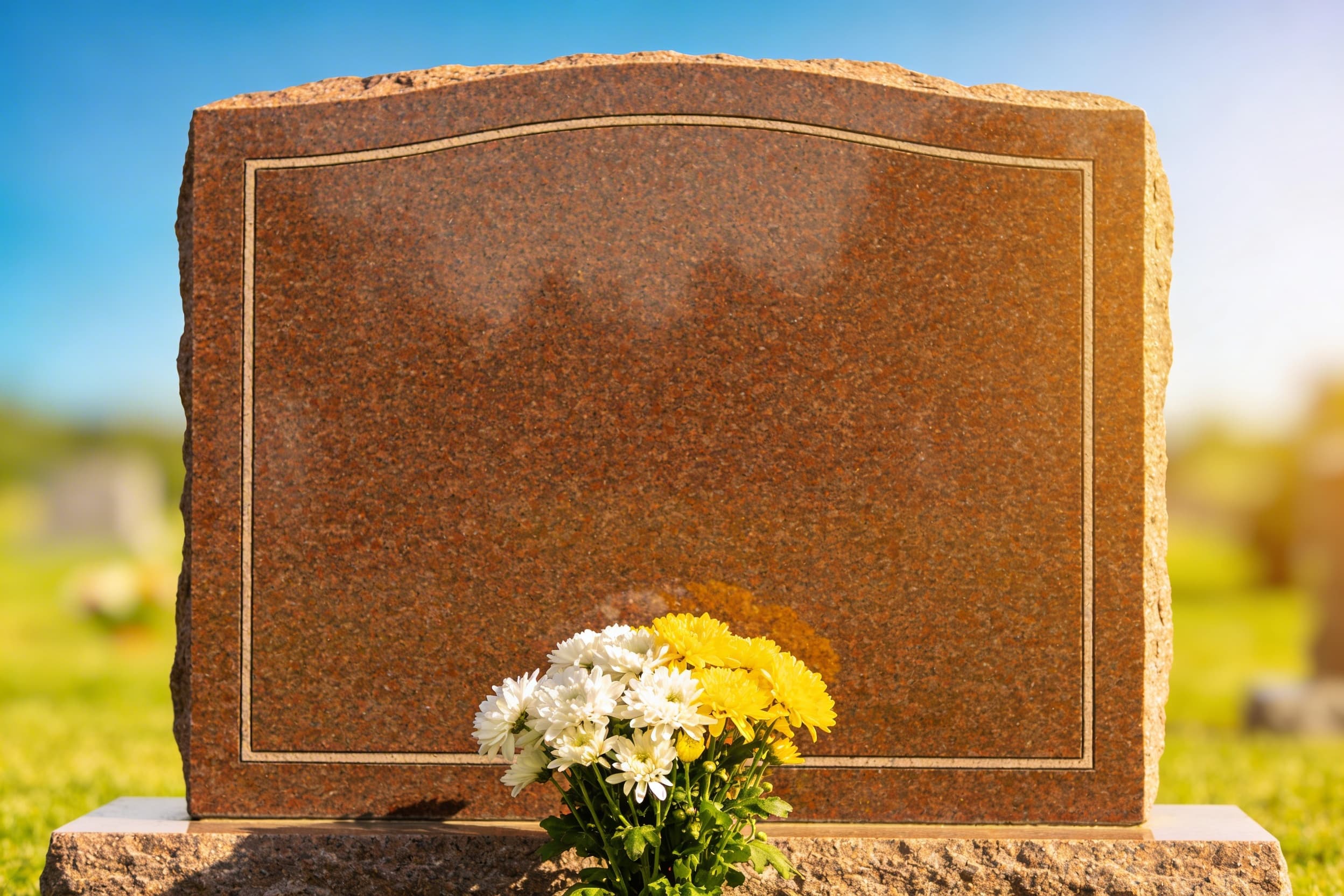 A clean, well-maintained granite headstone with fresh flowers at the base standing in a manicured cemetery on a sunny afternoon