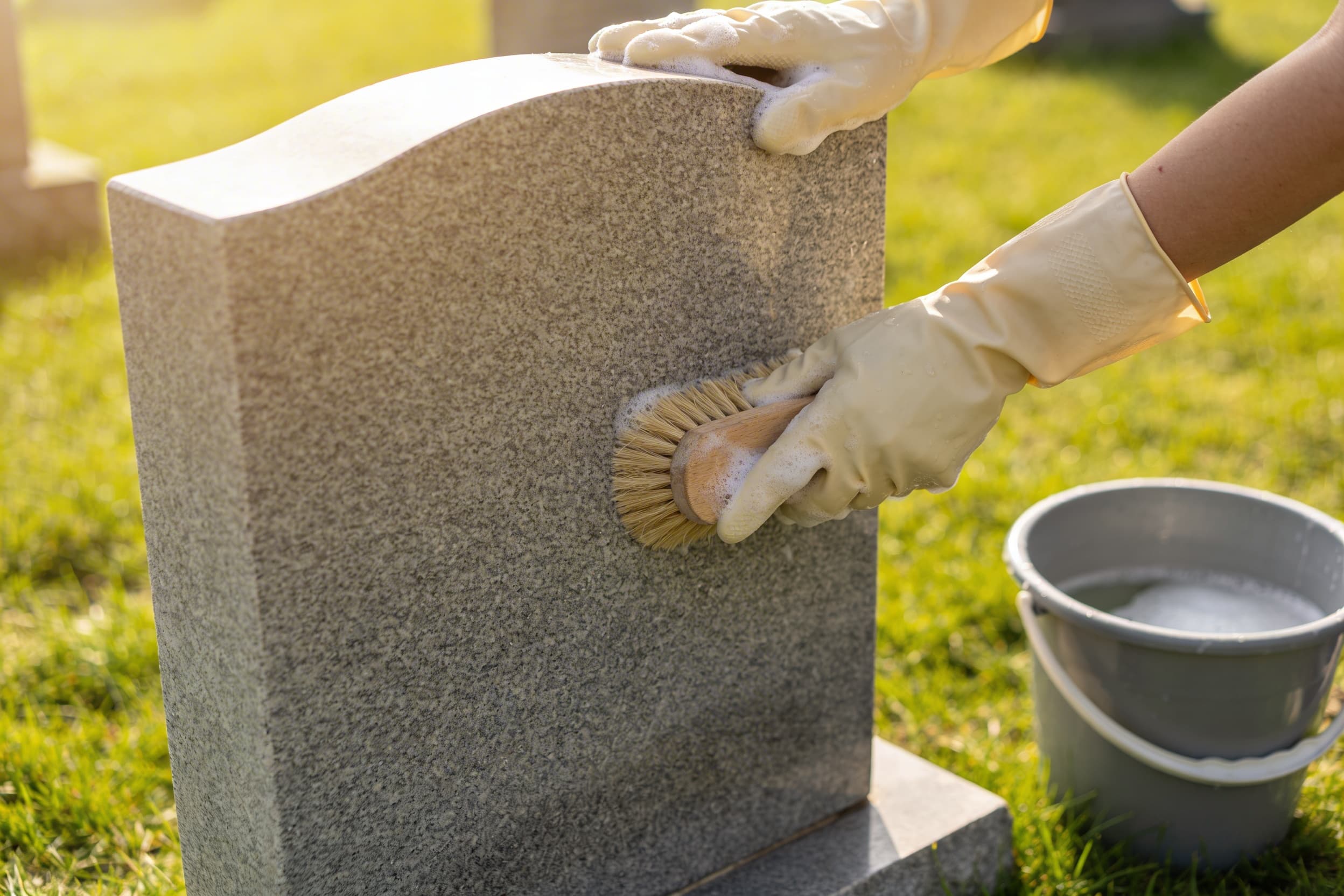 Hands wearing gloves gently scrubbing a granite headstone with a soft-bristle brush and soapy water