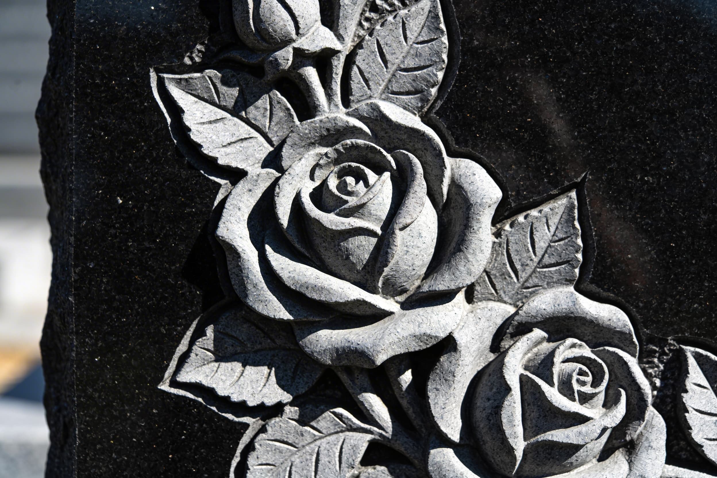 Close-up detail of hand-carved roses emerging from the surface of a polished black granite headstone, showing depth and shadow in the sculptural relief