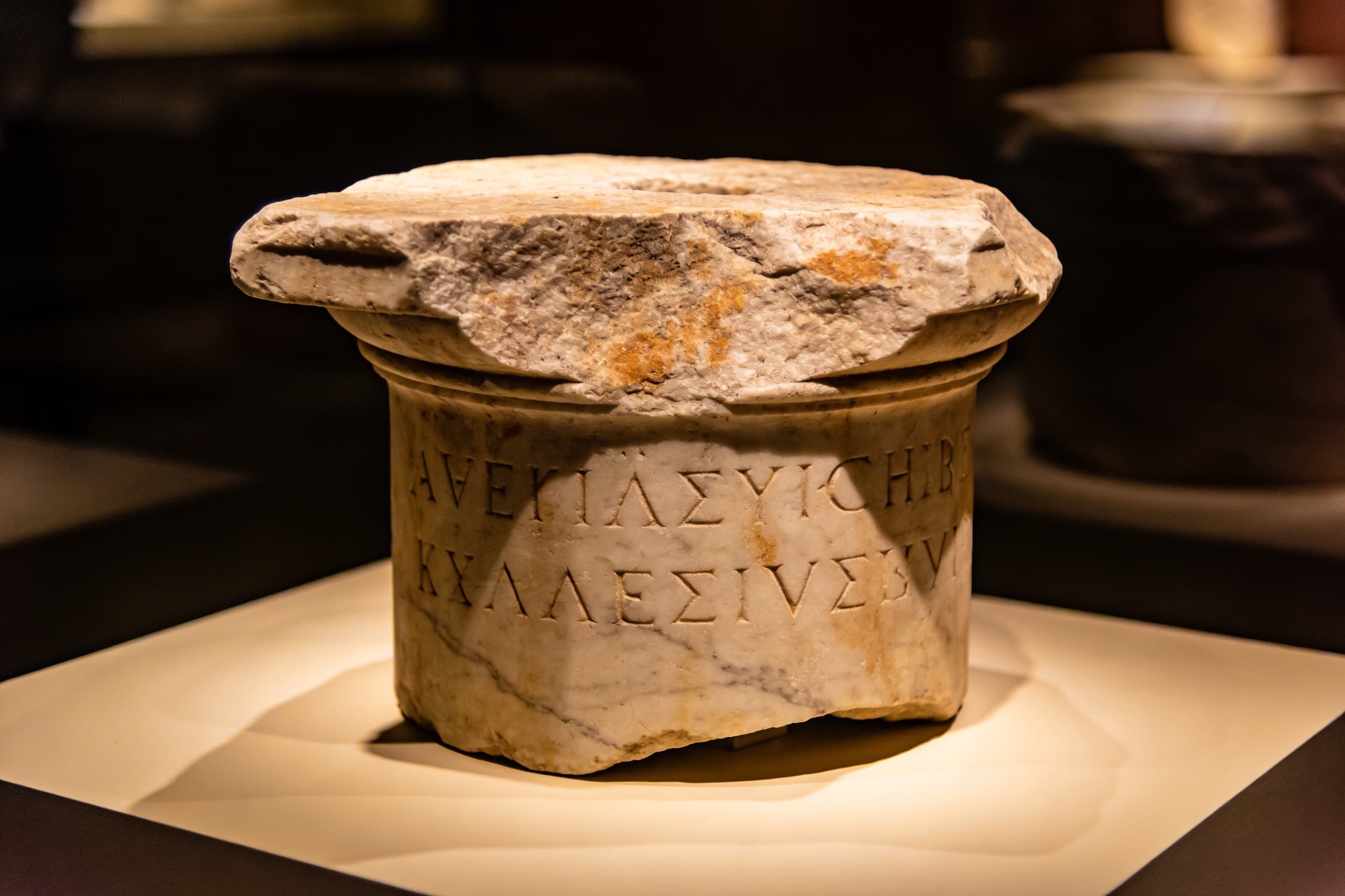 A classical stone column with faintly visible engraved text in an ancient cemetery setting surrounded by Mediterranean vegetation