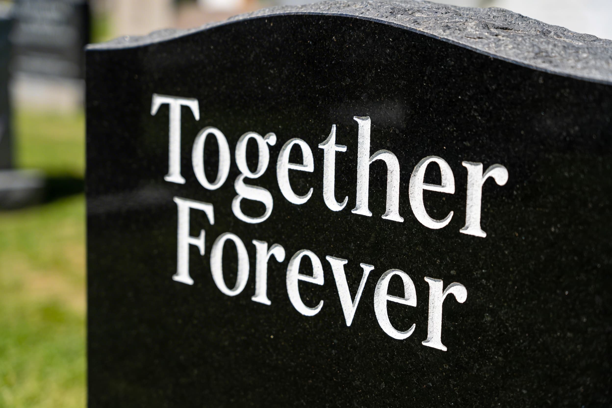 A close-up of a dark granite companion headstone with engraved text reading Together Forever in an elegant serif font with soft natural lighting