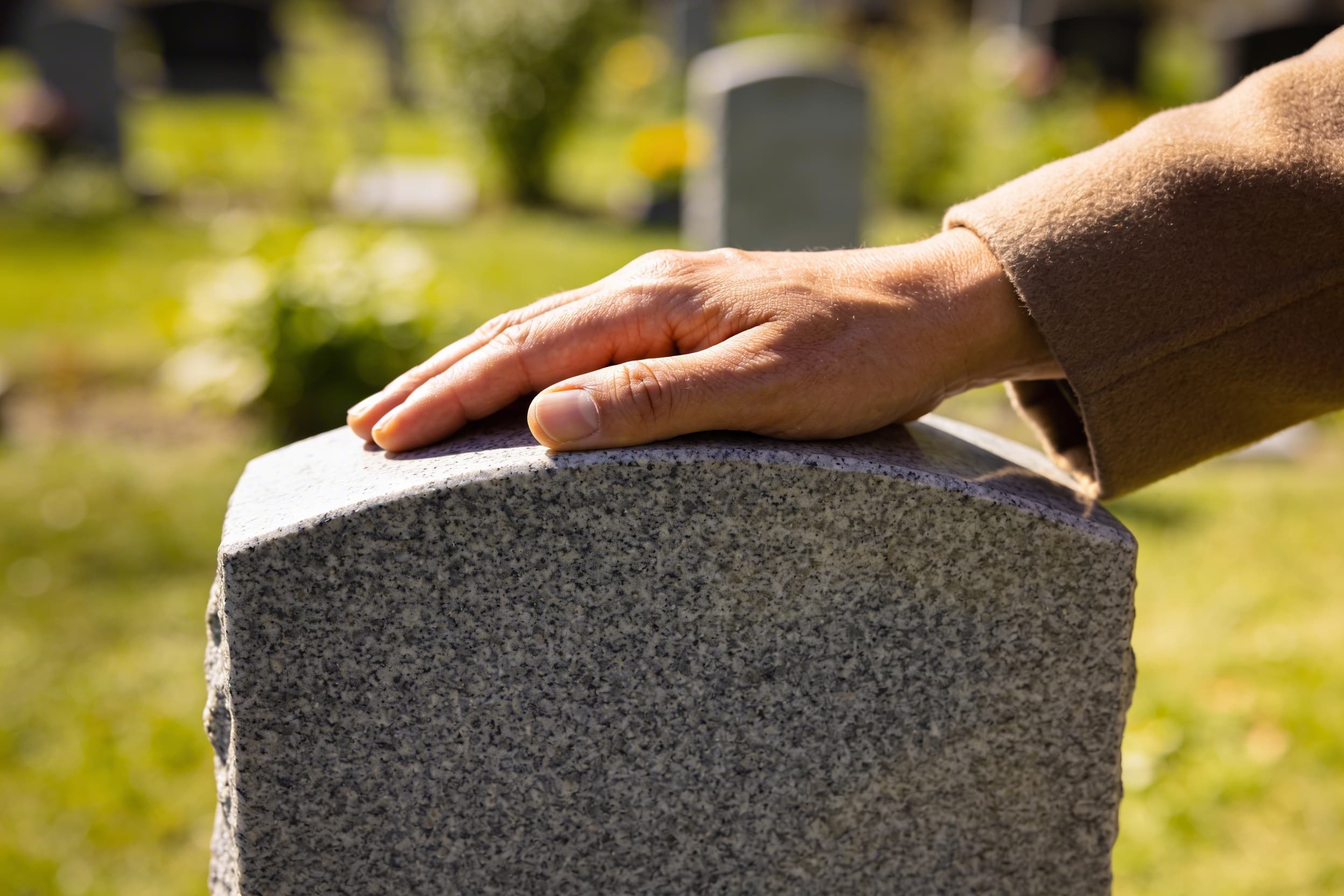A parent's hands gently resting on a small granite headstone with engraved letters in a quiet cemetery garden