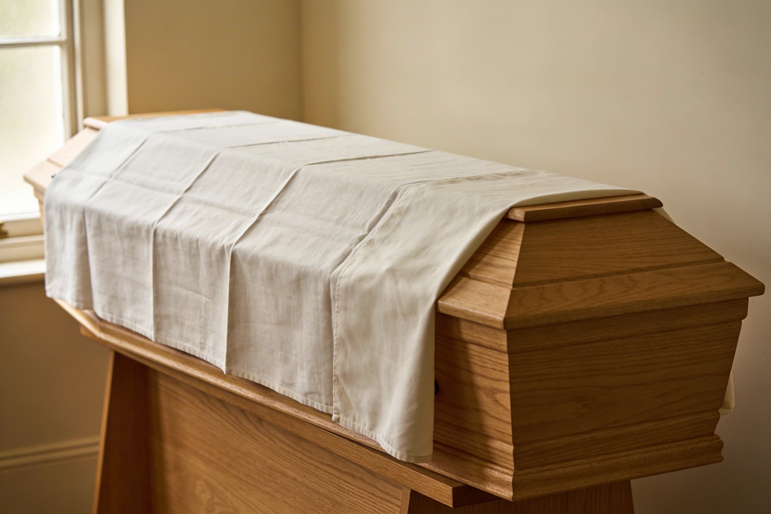 A simple unfinished wooden casket draped with a plain white linen shroud in a quiet, naturally lit room