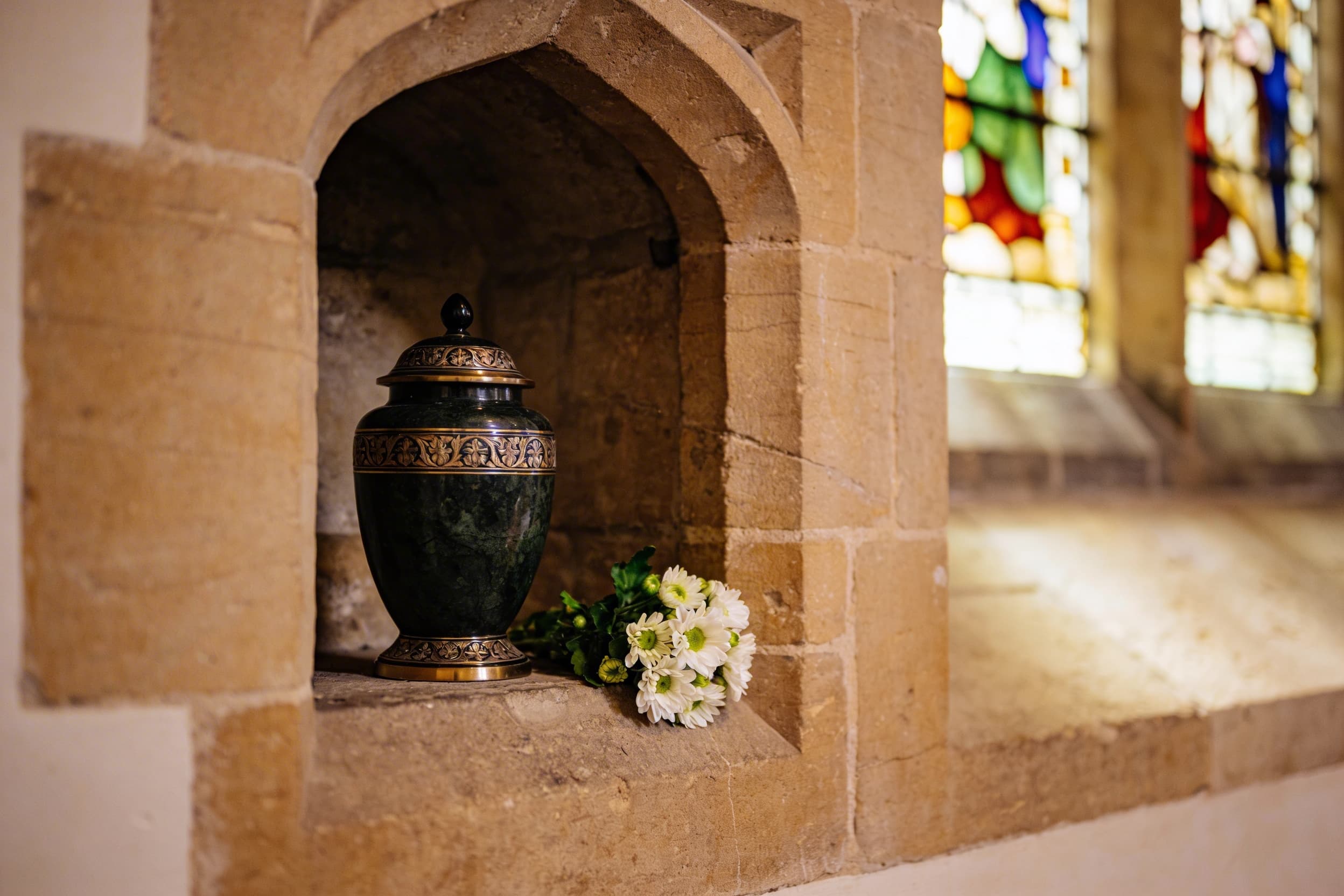 A columbarium niche inside a Catholic church with a small urn and fresh flowers placed respectfully beside a nameplate