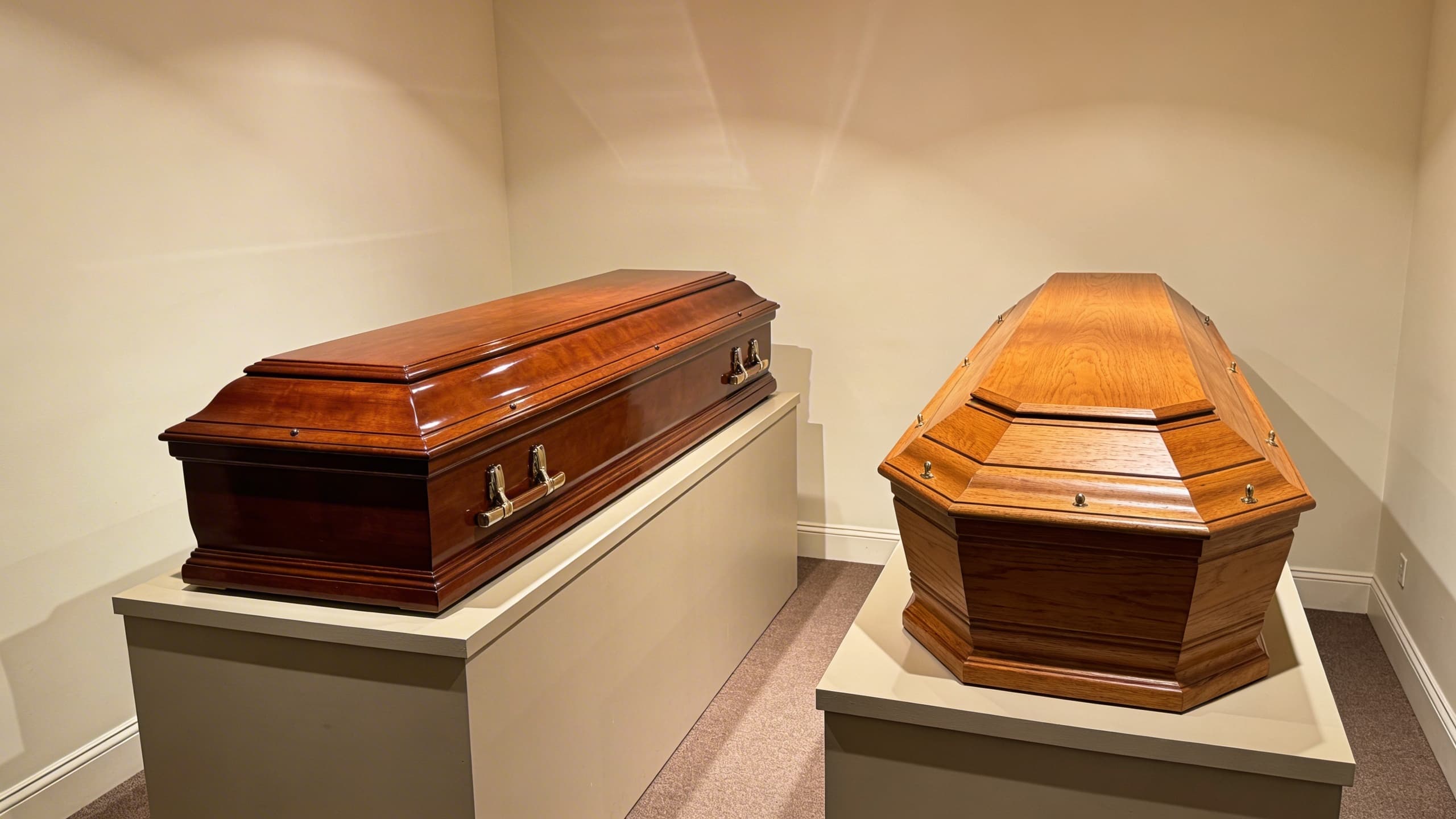 A rectangular four-sided casket next to a tapered six-sided coffin displayed in a funeral showroom with soft lighting