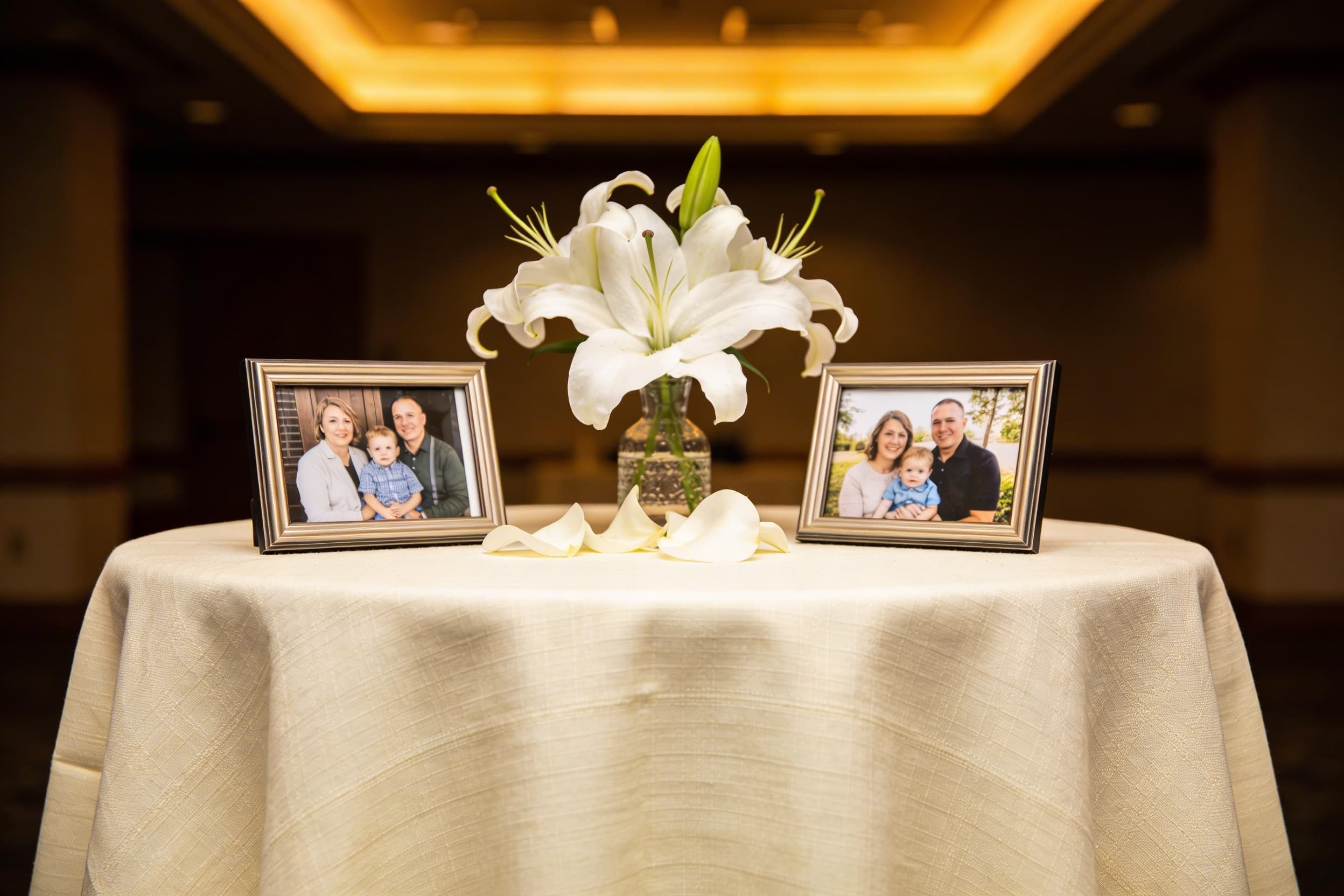 Two framed family photographs and a vase of white lilies arranged on a cream linen cloth at a memorial service with warm golden lighting