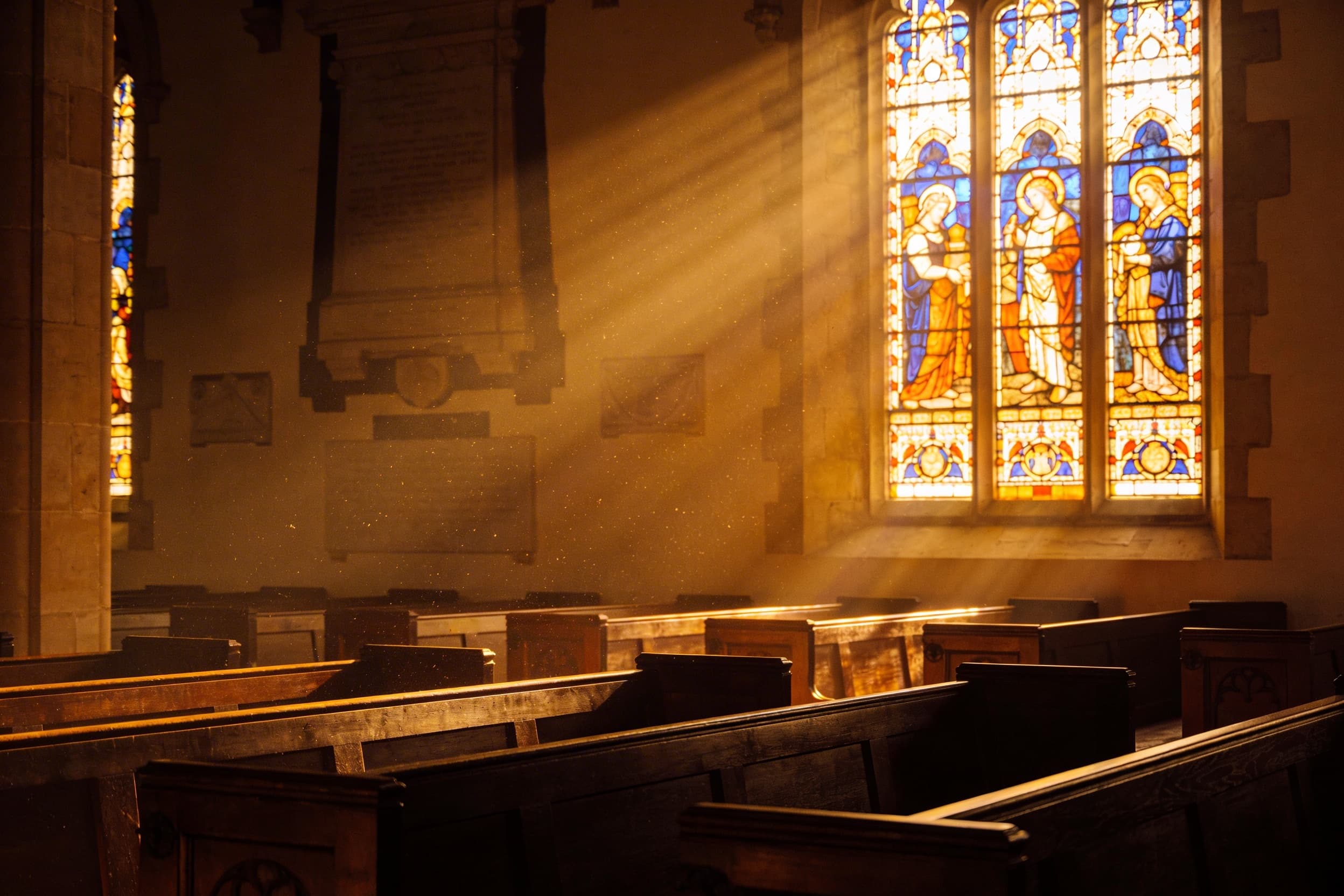 Warm light streaming through a stained glass church window illuminating wooden pews