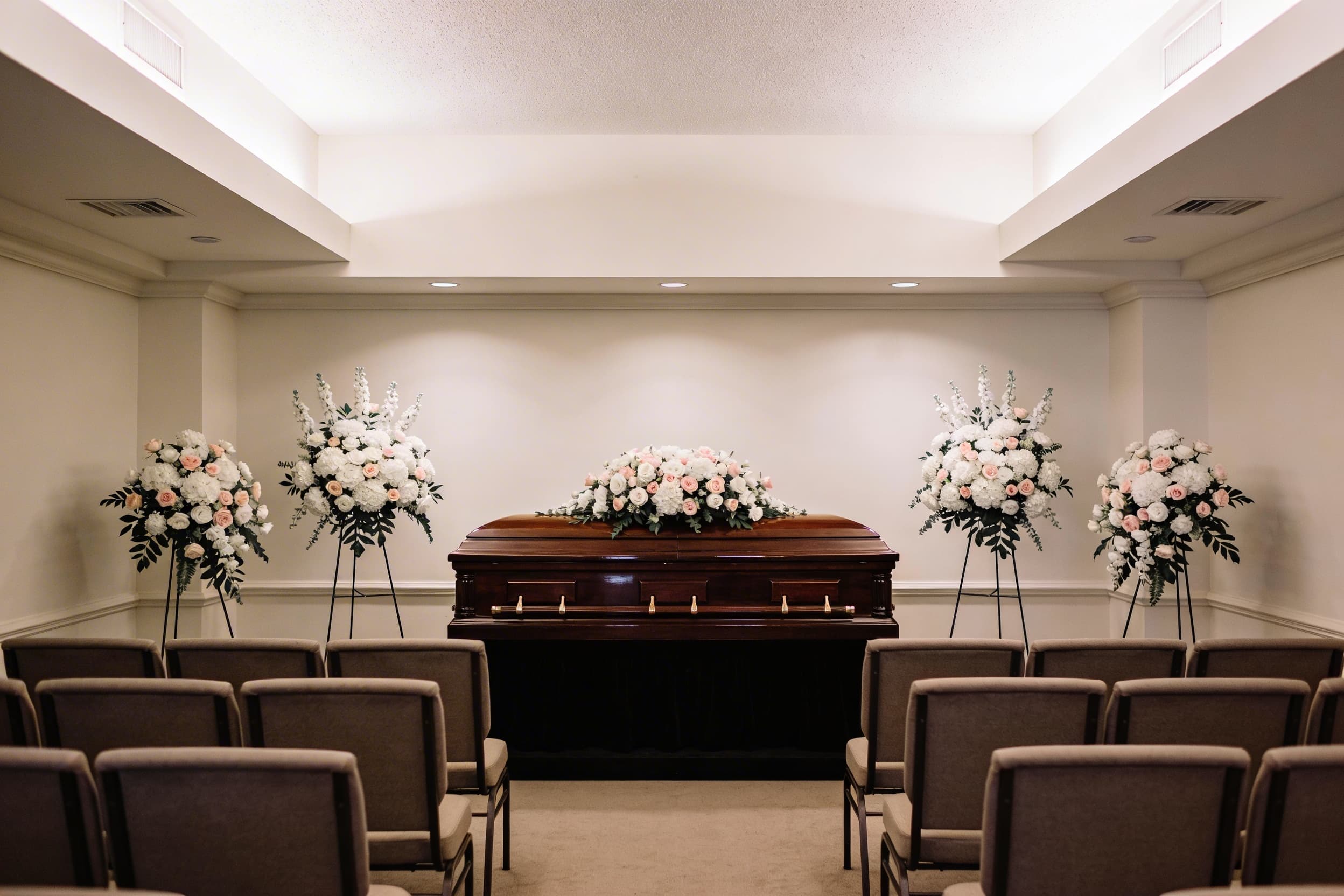 A funeral home viewing room with a polished dark wood casket surrounded by white and pink floral arrangements and upholstered seating
