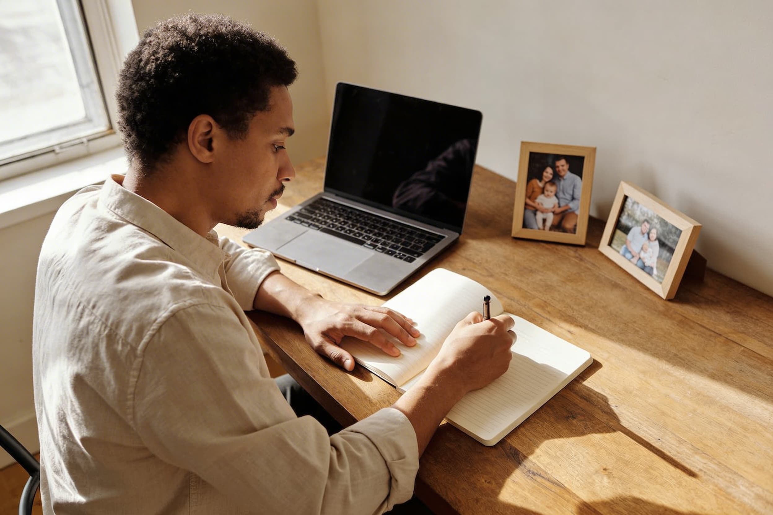A person sitting at a desk writing an obituary with family photographs and a notebook beside the laptop
