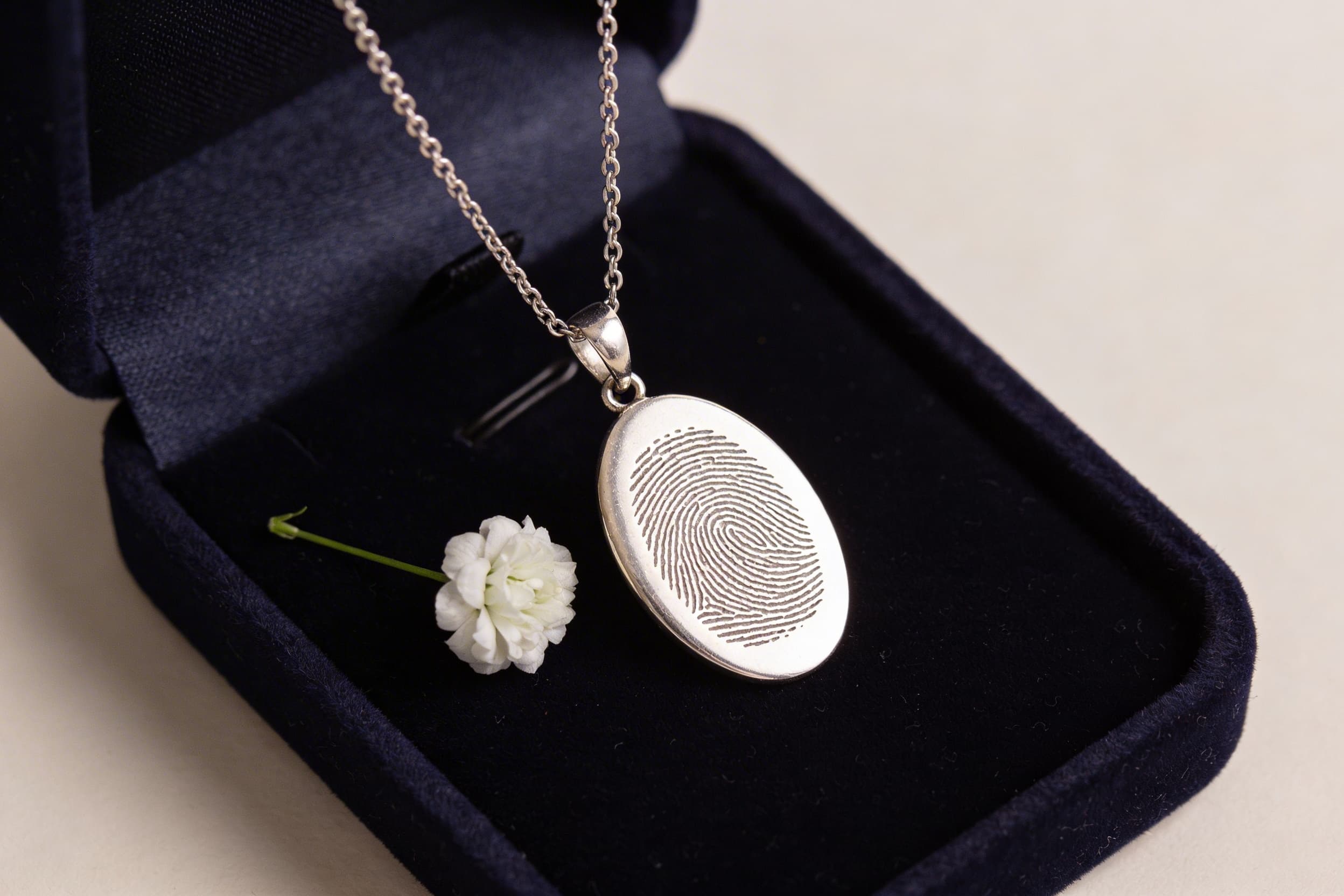 A silver fingerprint memorial pendant necklace displayed on dark velvet next to a small white flower