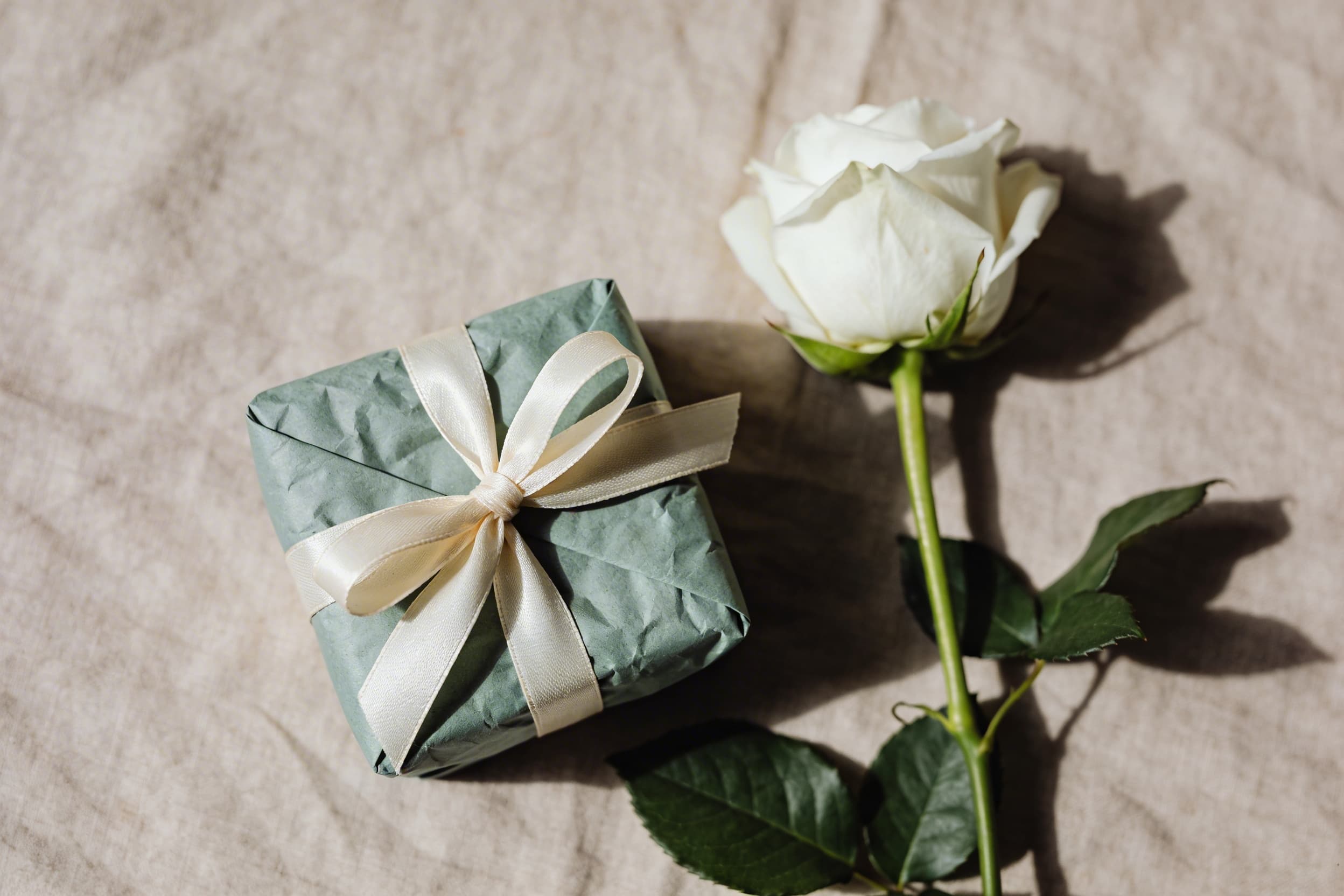 A small wrapped memorial keepsake gift in sage green tissue paper tied with cream ribbon beside a single white rose on a linen surface