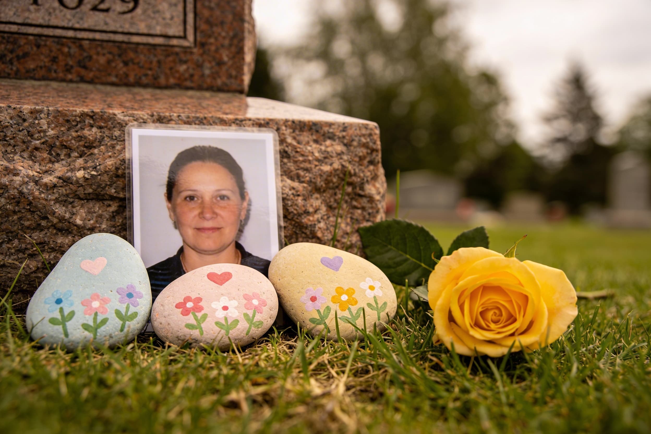 Handmade grave decorations including painted river rocks with hearts and flowers arranged beside a granite headstone with a fresh yellow rose