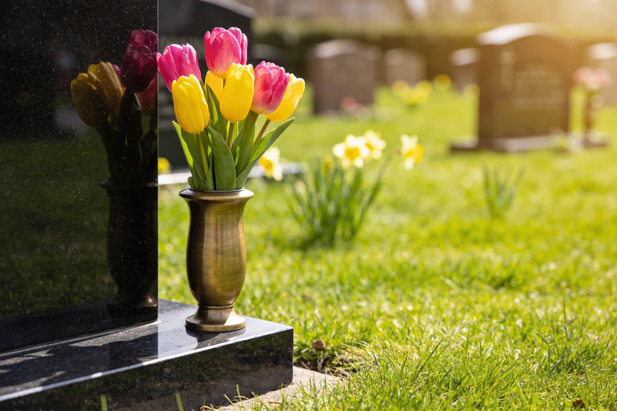 A small weatherproof vase of bright tulips placed beside a granite headstone in a green cemetery during spring
