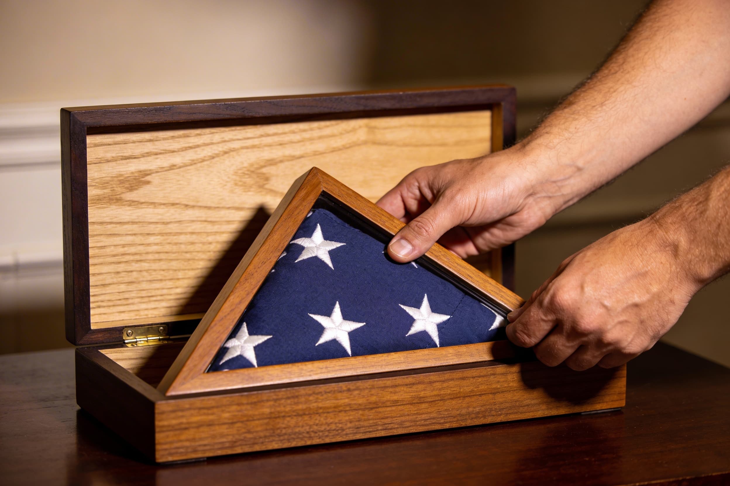 A pair of hands carefully placing a folded American burial flag into an oak flag display case from the rear opening