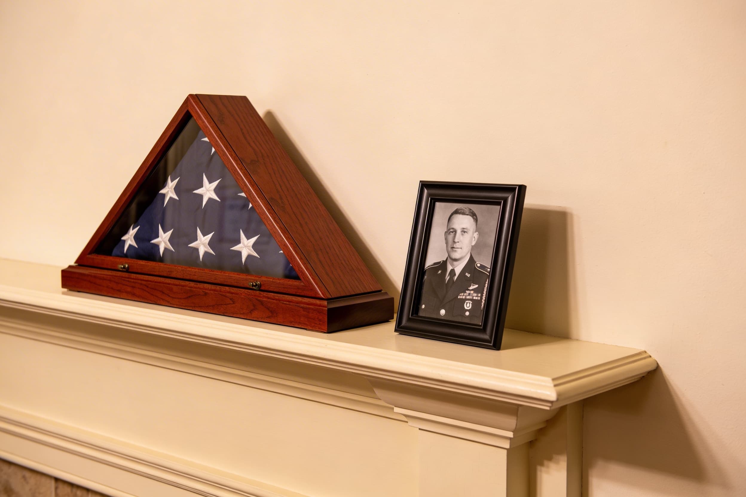 A folded American burial flag displayed in a cherry wood flag case on a home mantel beside a framed military portrait