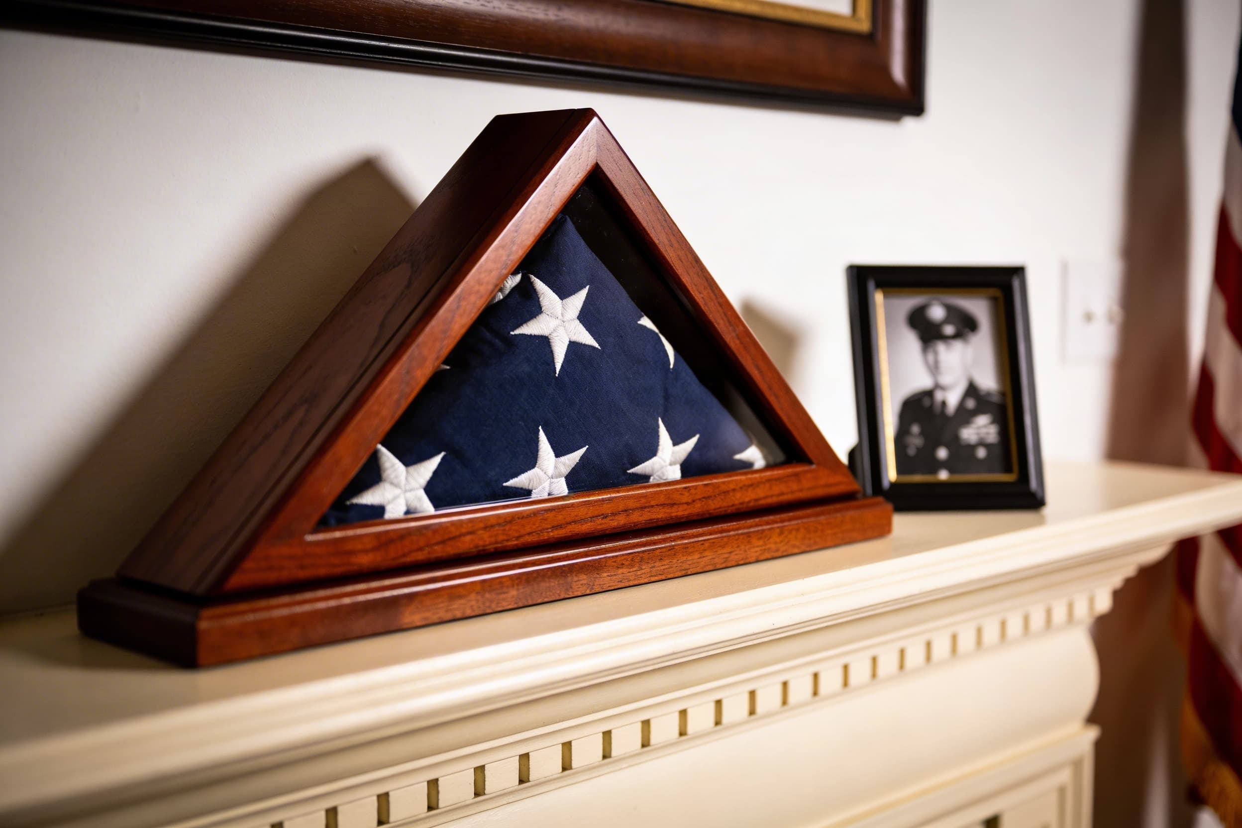 A triangular flag display case with a folded American flag on a fireplace mantel next to a framed military portrait