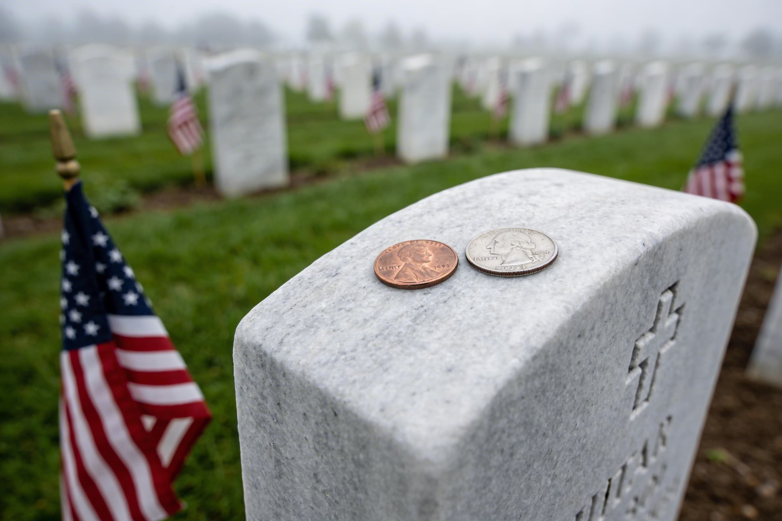 A penny and quarter placed on a veteran's granite headstone with small American flags beside it in a veterans cemetery