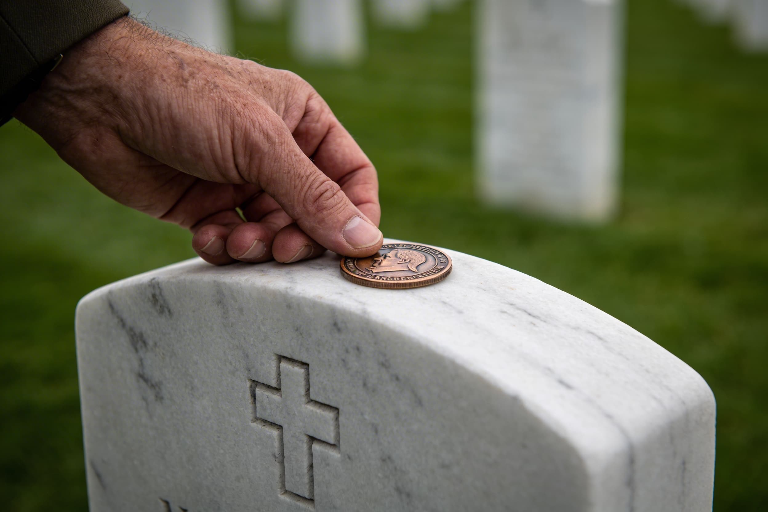 A veteran's hand gently placing a unit challenge coin on top of a fellow service member's white marble headstone in a peaceful cemetery setting