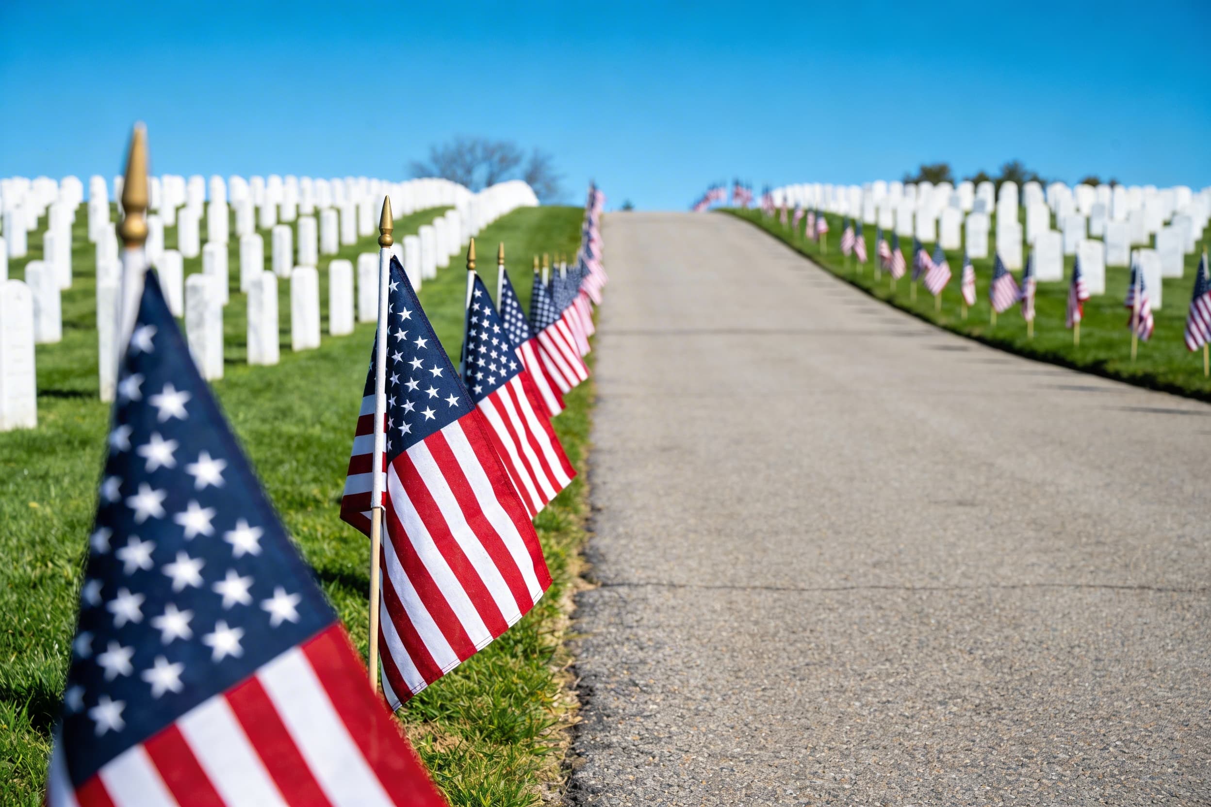 Rows of American flags lining the Avenue of Flags at a national cemetery on a patriotic holiday with green grass and headstones in the background